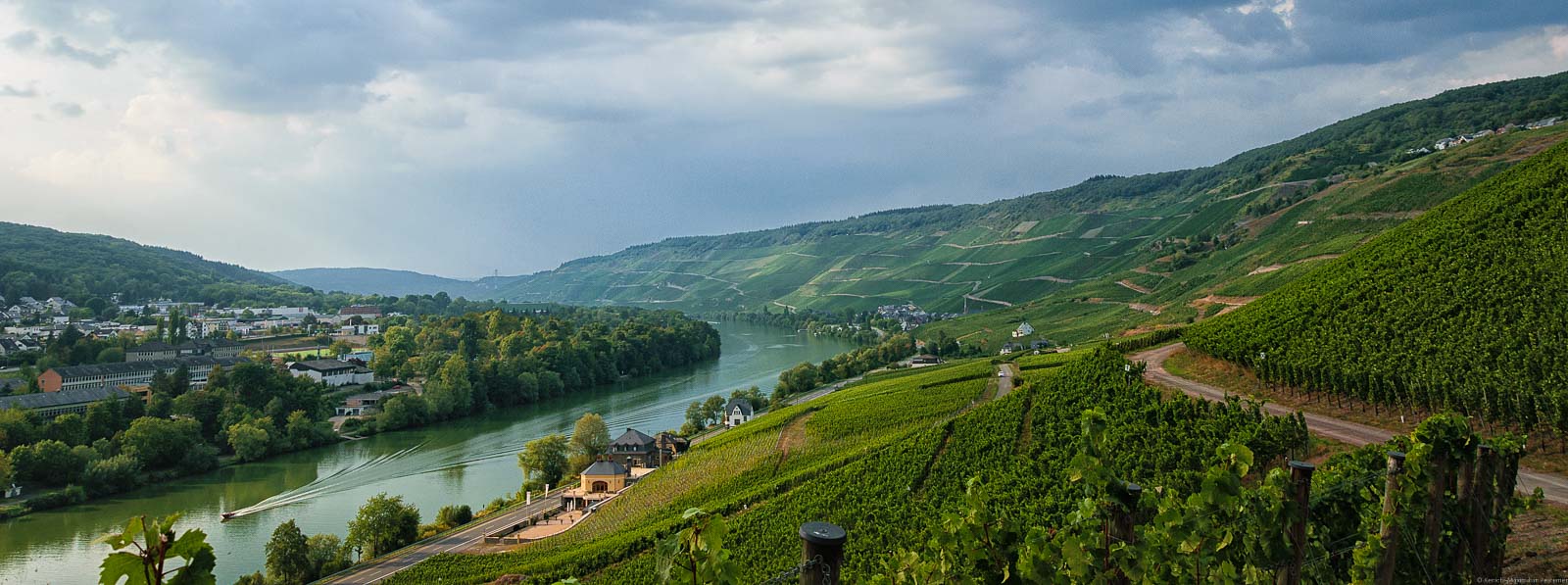 Vorne rechts befindet sich der Weinberg Bernkasteler Lay. Dort stehen vor allem Riesling-Reben. Links erkennt man der Verlauf des Flusses Mosel. Auf der Mosel fährt ein Boot. Im Hintergrund sind weitere Weinberge und Wolken am Himmel