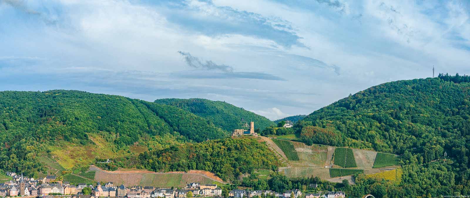Am unteren Rand liegt der Ort Bernkastel-Kues. Über dem Ort liegen Weinberge, Wälder und Himmel. In der Mitte befindet sich die Burg Landshut. Unterhalb der Burg Landshut liegt der Bernkasteler Schloßberg.
