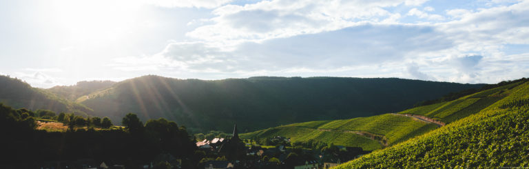 Sonnenuntergang an der Mosel. Links vorne ist ein Felsen. In der Mitte im Tal ist der Ort Neumagen-Dhron mit einer Kirche. Rechts liegt der Weinberg Dhroner Hofberger. Oben sind große Wolken.