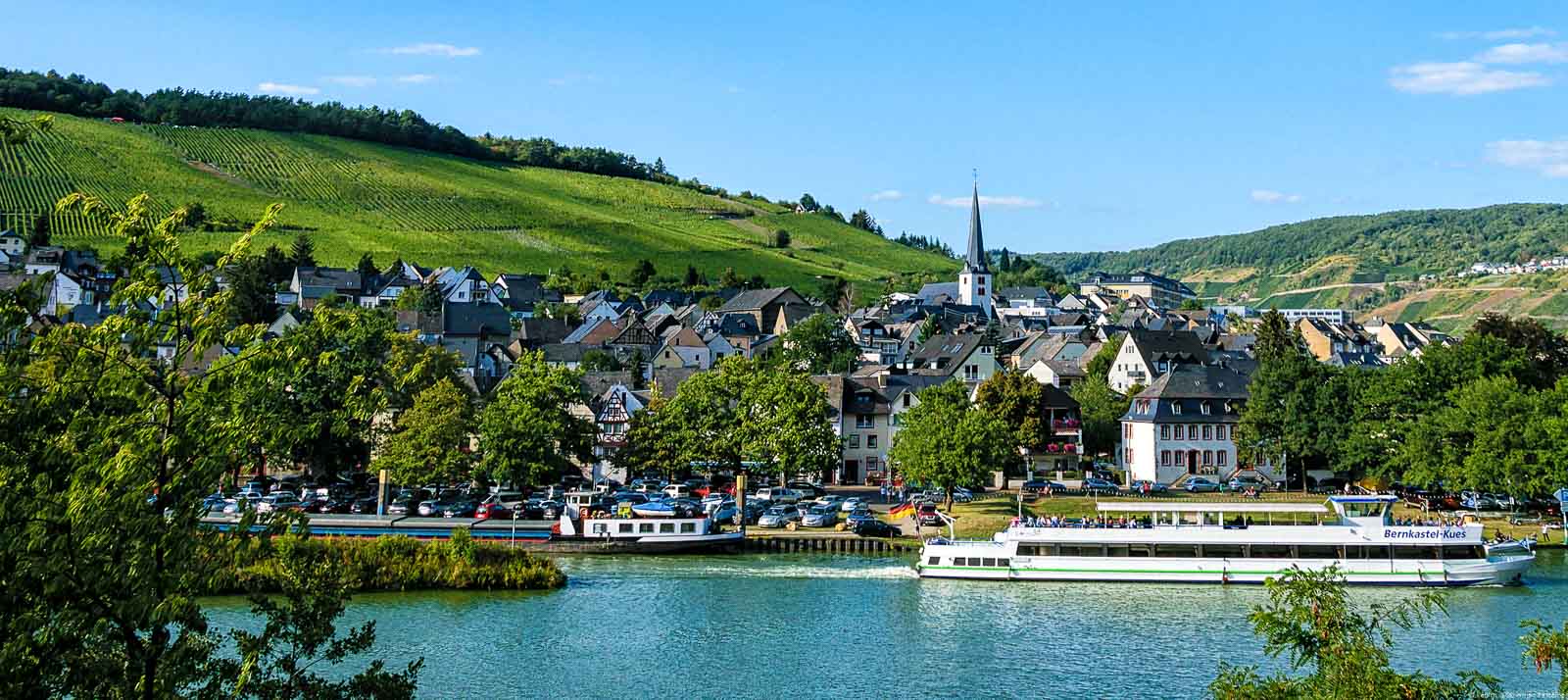 Vor fließt die Mosel. Am Ufer gegenüber liegt der Ort Bernkastel-Kues. Hinter dem Ort liegt der Weinberg.