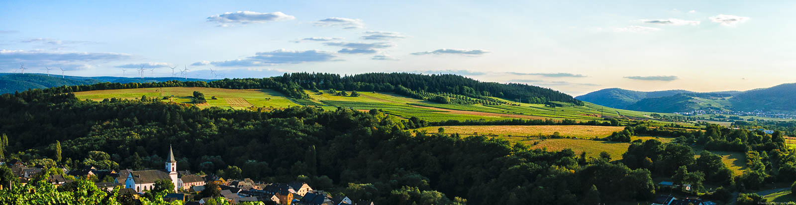 Die Lambertuslay liegt auf dem Rücken der Brauneberger Juffer. Vorne links erkennt man den Ort Maring-Noviand. Im Hintergrund liegt das Dorf Osann-Monzel. Am leicht blauen Himmel bewegen sich einige Wolken. Im Hintergrund sind bewaldete Hügel.