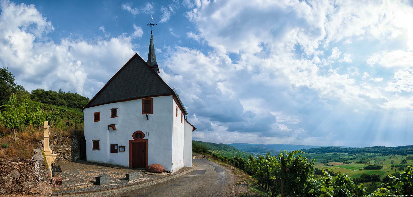 Auf dem Schlossberg bei Lieser befindet sich eine alte Kapelle. Unten breitet sich die Ebene vor dem Dorf aus. Am Himmel sind Wolken aus den Sonnenstrahlen auf den Boden strahlen. Der Weinberg liegt im Bereich der Mittelmosel.