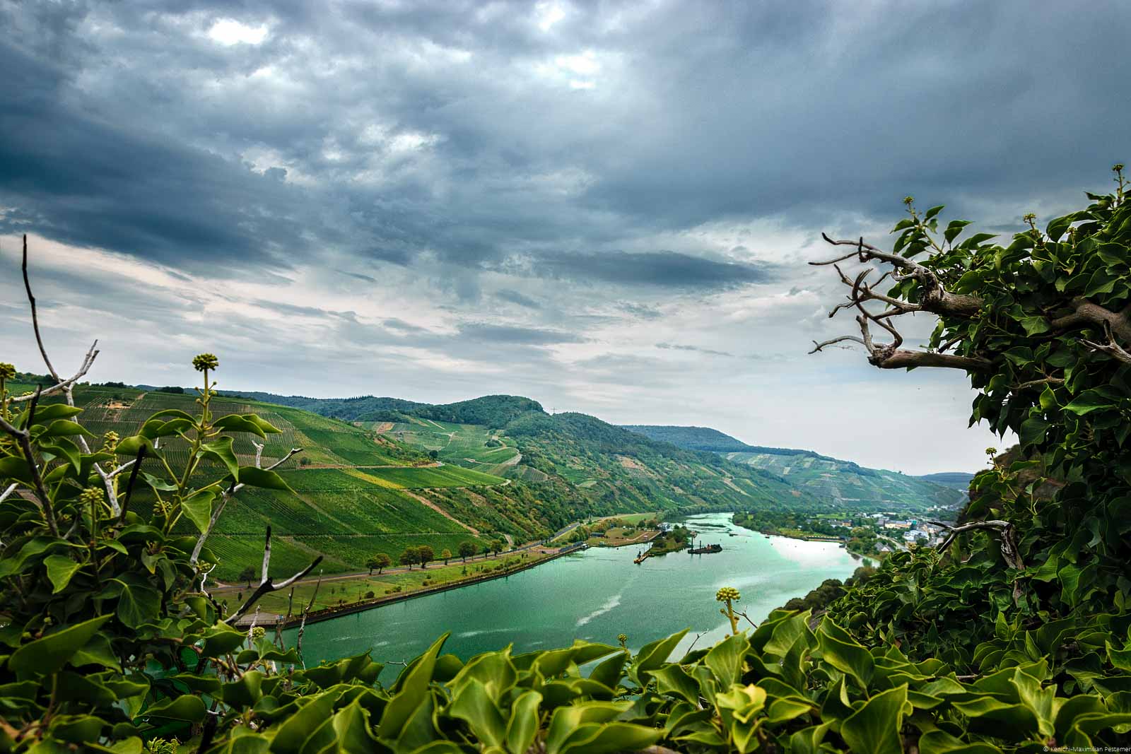Vorne links am Ufer der Mosel liegt das Minheimer Kapellchen am Ufer der Mosel. Der Fluss fließt in der Mitte des Bildes bei einer Stausperre kurz vor Wintrich. Der Himmel ist stark bewökt.