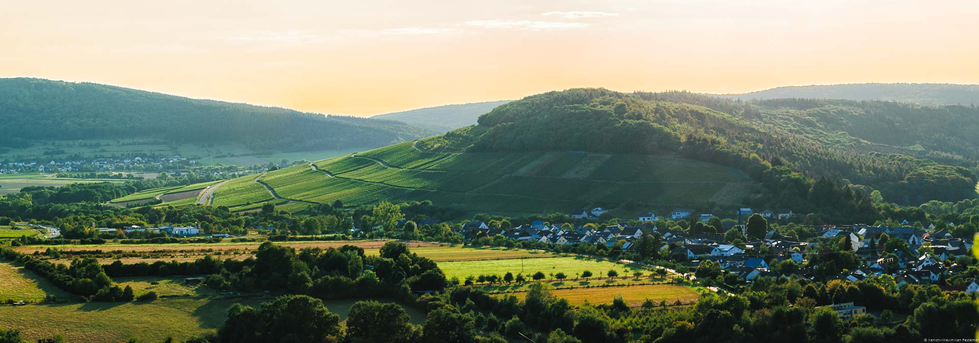 Vorne sind Wälder und Felder. Vorne rechts liegt der Ort Maring-Noviand. Dahinter erhebt sich der steile Weinberg Osanner Rosenberg beim Sonnenuntergang. Im Hintergrund liegt der Ort Osann-Monzel mit bewaldeten Hügeln.