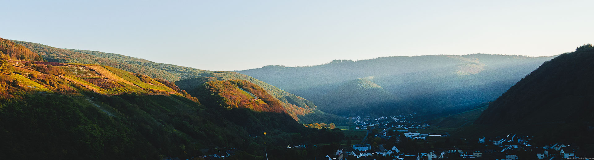 Sonnenstrahlen strahlen Abends seitlich in das Ahrtal auf den Weinberg Dernauer Goldkaul. Im Hintergrund erblickt man den Ort Rech. Die Hügel an beiden Seiten des Tales sind bewaldet.