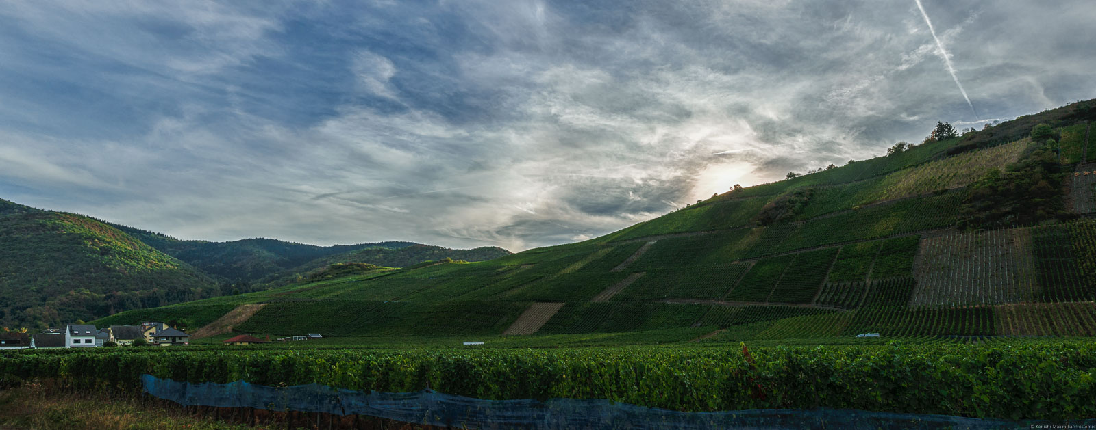 Vorne liegt der flache Weinberg Dernauer Burggarten. In der Mitte liegt der steile Weinberg Recher Herrenberg. Links auf dem Bild sind Häuser. Oben geht die Sonne am Weinberg unter. Der Himmel ist bewölkt.