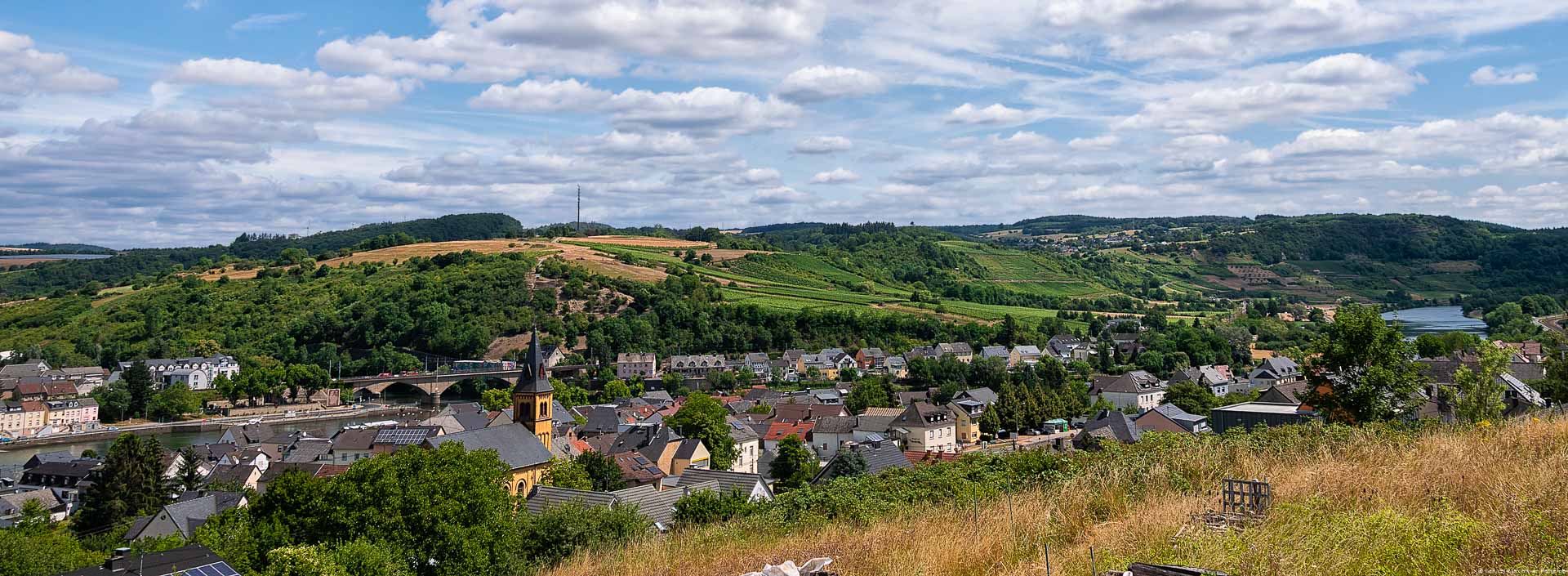 Blick auf Weinberge wie zum Beispiel der Langsurer Brüderberg an der Obermosel. Vorne im Tal fließt der Fluss Mosel. Am gegenüberliegenden Ufer der Mosel liegt der Ort Wasserbillig. Am blauen Himmel sind viele Wolken.