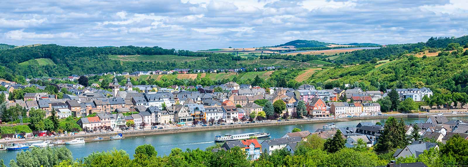Im Vordergrund fließt die Mosel. Am anderen Ufer liegt der Ort Luxemburg. Im Hintergrund liegen Weinberge. Man erkennt hinter dem Ort Wasserbillig den Langsurer Brüderberg am Ufer des Flusses Sauer. Der Himmel ist bewölkt.