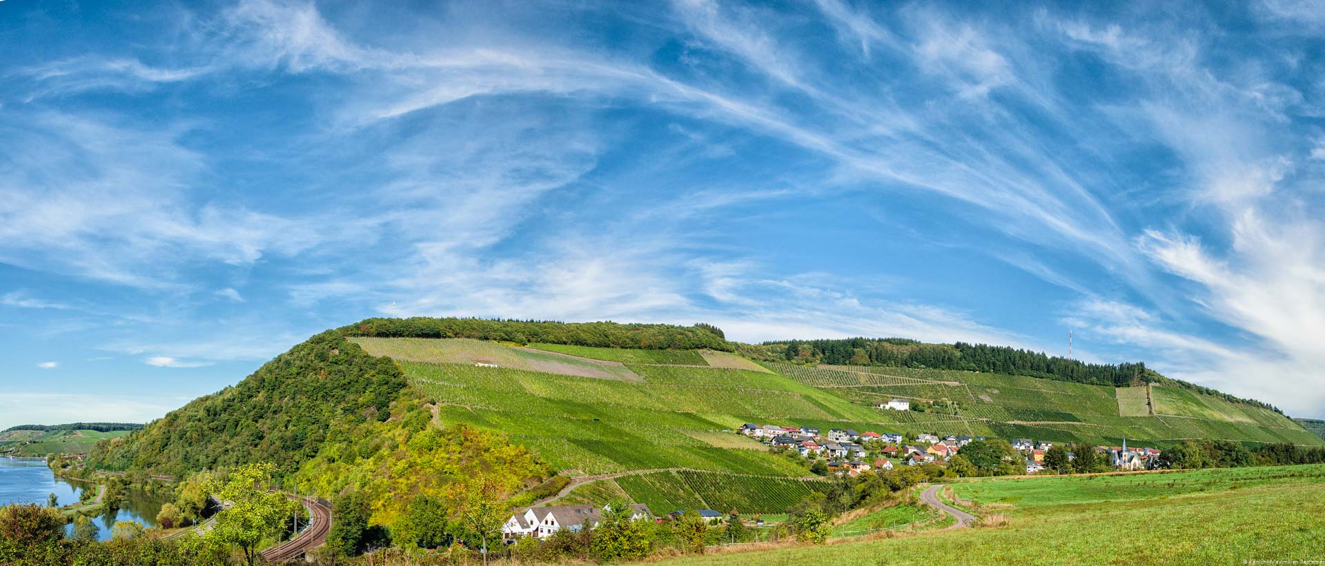 Links fließt die Saar. In der Mitte befindet sich der steile Weinberg Ockfener Bockstein. Oben ist ein blauer Himmel mit einigen Wolken.