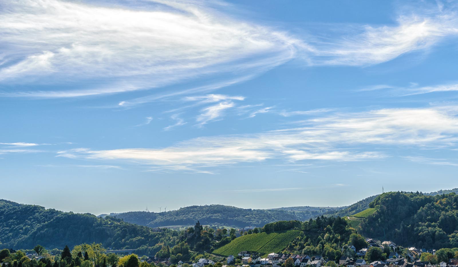 Aussicht auf den Ort Saarburg, den Fluss und die Weinberge. Man erkennt rechts den Ort Niederleuken. In der Mitte erkennt man die Saarburg und den Weinberg Saarburger Schlossberg daneben. Im Hintergrund sind bewaldete Hügel. Am blauem Himmel fliegen einige weiße Wolken.