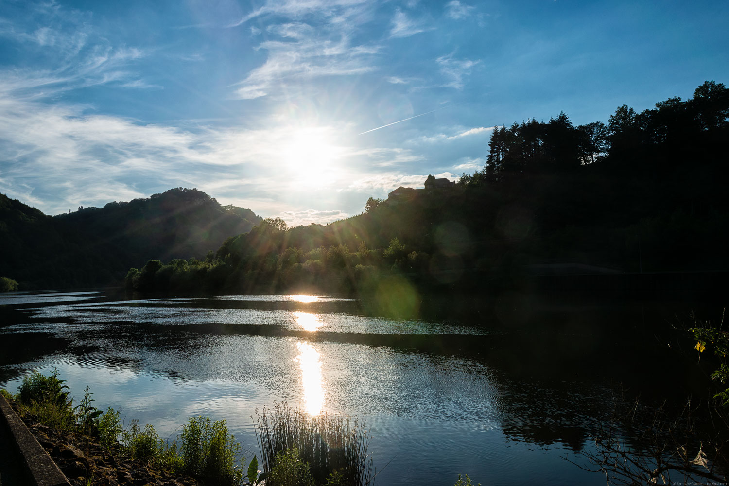 Vorne fließt der Fluss Saar. In der Saar erkennt man die Reflexionen der Sonne. Am Ufer gegenüber befindet sich Wald, der Weinberg Serriger Würtzberg und ein Weigut. Es ist dunkel und die Sonne geht am leicht bewölkten blauen Himmel unter.de