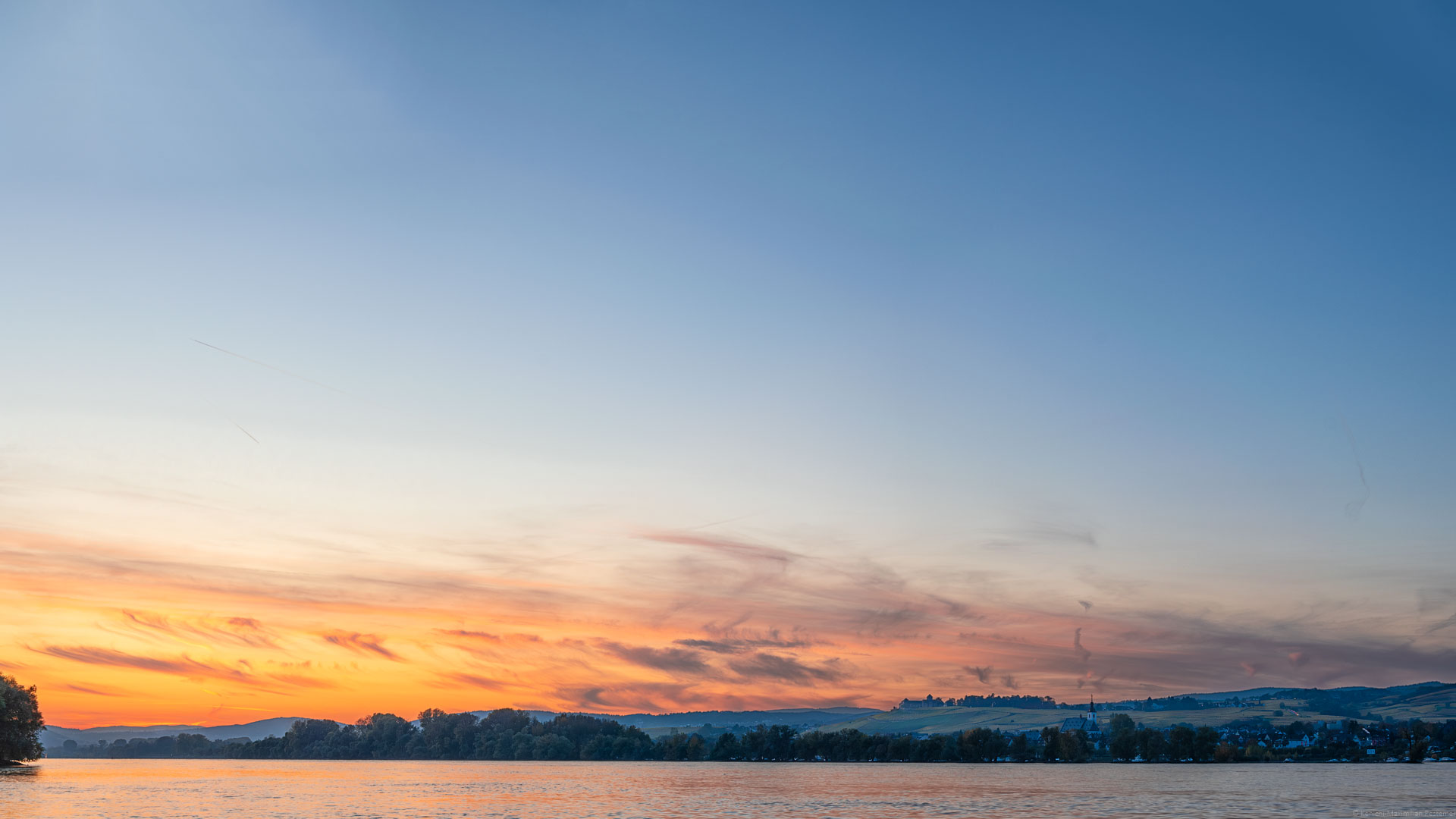 Feuriger Sonnenuntergang bei Oestrich-Winkel am Rheingau. Vorne unten im Bild verläuft der Fluss Rhein. Am anderen Ufer erkennt man Bäume, Häuser und Weinberge des Rheingaus. Im Hintergrund erblickt man das Schloss Johannisberg- Der Himmel ist orange, rot und blau.