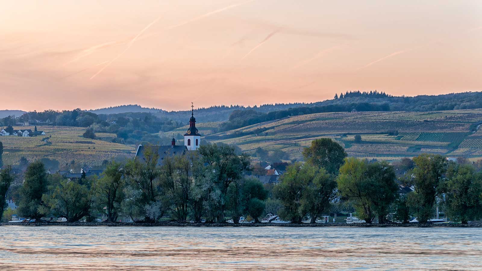 Von der gegenüberliegenden Seite des Rheins kann man feurige Sonnenuntergänge am Rheingau genießen. Vorne im unteren Teil des Bildes fließt der Rhein. Am Ufer gegenüber erkennt man die Kirche und die Häuser des Ortes Oestrich-Winkel. Hinter dem Ort liegen Weinberge mit Wäldern oberhalb. Der Himmel ist rötlich gefärbt.