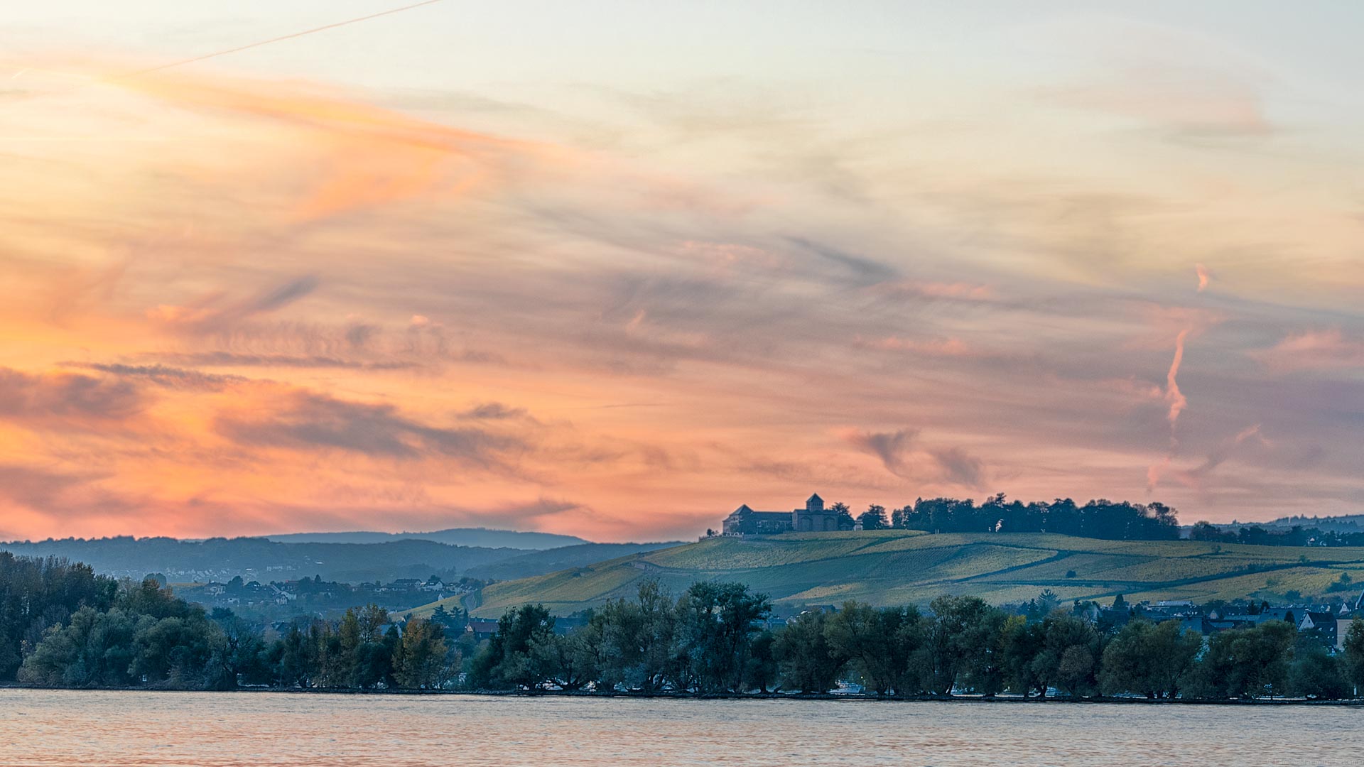 Im Vordergrund fließt der Fluss Rhein. Am Ufer mit Bäumen und Ort Oestrich-Weinkel, erkennt man den Weinberg Schloss Johannisberg mit herbstlicher Färbung auf dem gleichnamigen Weinberg. Man erblickt einige Häuser und Gebäude auf den Weinbergen. Diese wurden von den Zisterzienser-Mönchen erbaut. Der Himmel oben ist orange und bläulich gefärbt.