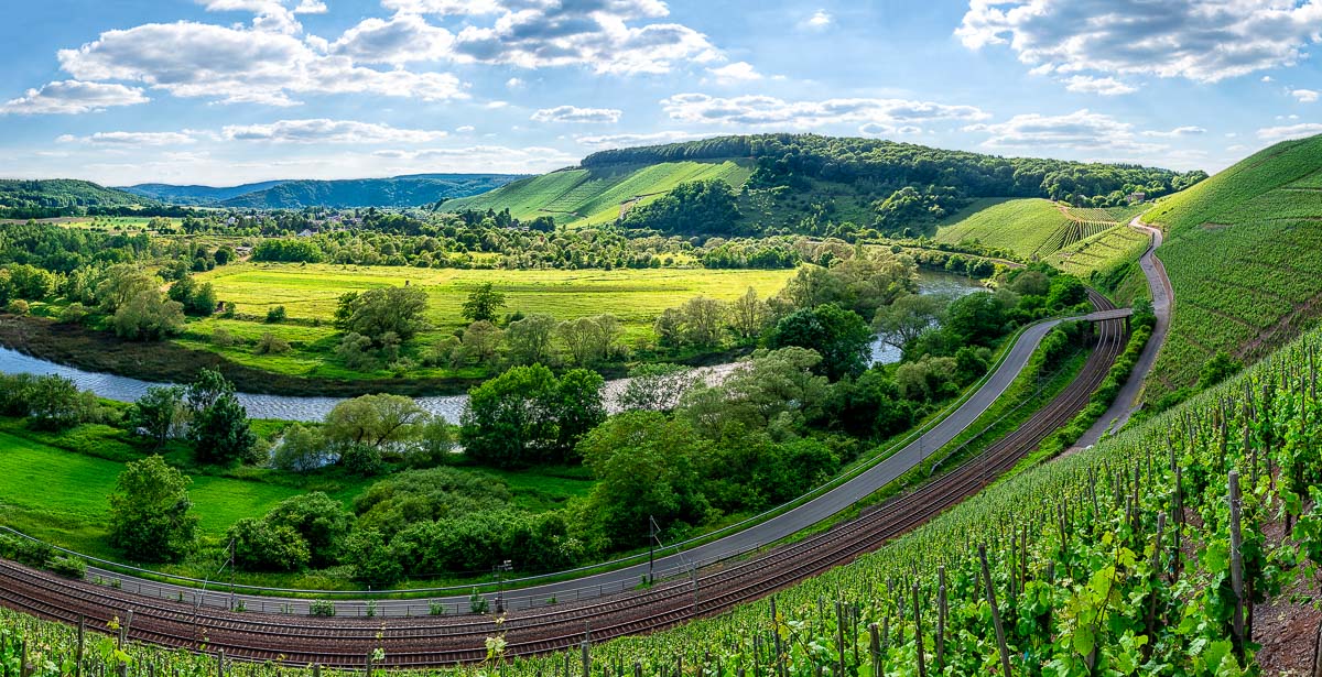 Mitten im Bild biegt sich die Saarschleife. Vorne sind Weinberge und Gleise sowie eine Straße. Am anderen Ufer sind Wiesen, Wälder und der Ort Kanzem. Im Hintergrund liegen weitere Weinberge sowie bewaldete Hügel. Der Himmel ist blau und etwas bewölkt.