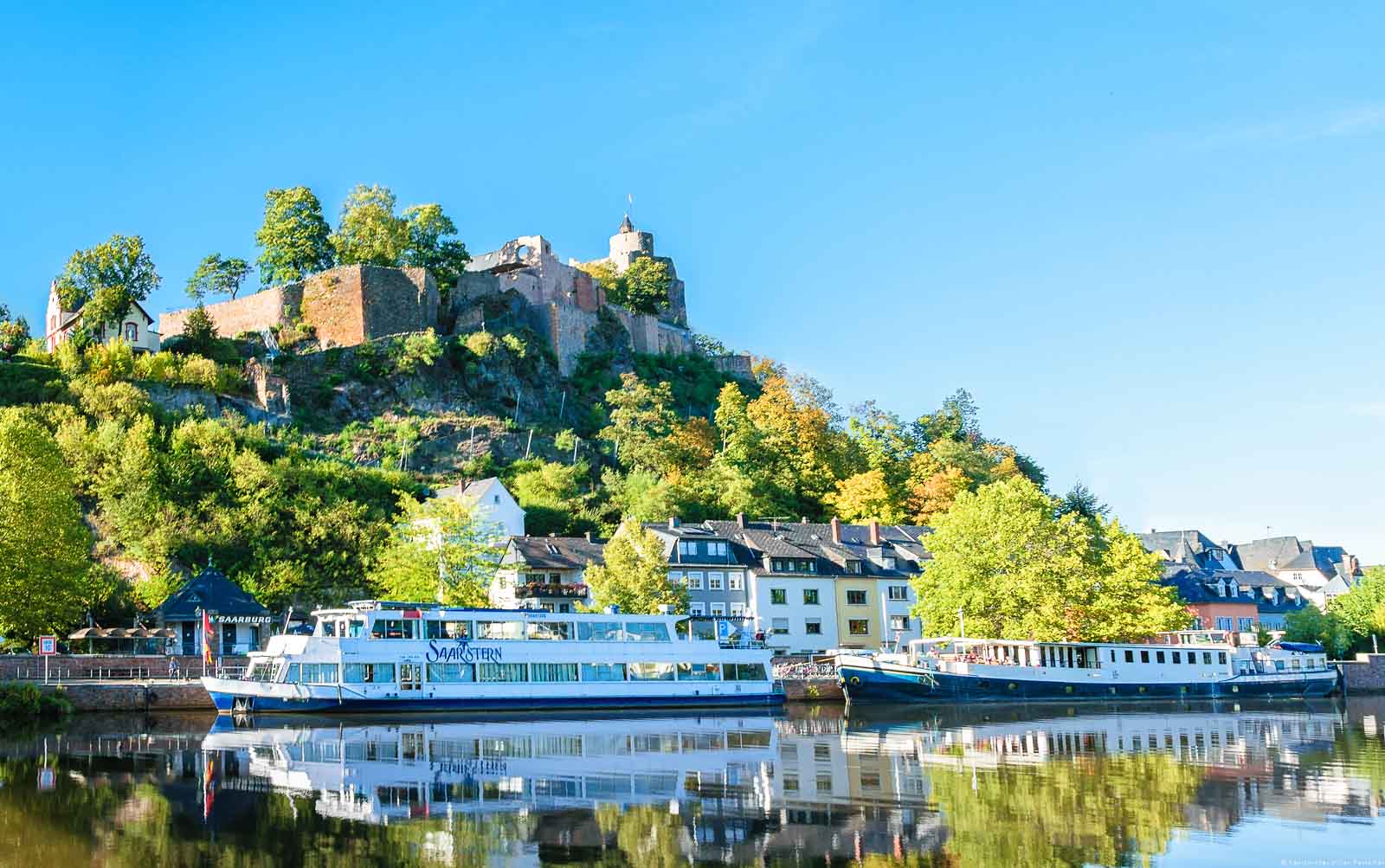 Vorne fließt der Fluss Saar. Am anderen Ufer ist ein Schiff. Dahinter befinden sich Häuser der Stadt Saarburg und ein Schloss auf einem steilen bewaldeten Hügel. Der Himmel ist oben blau.