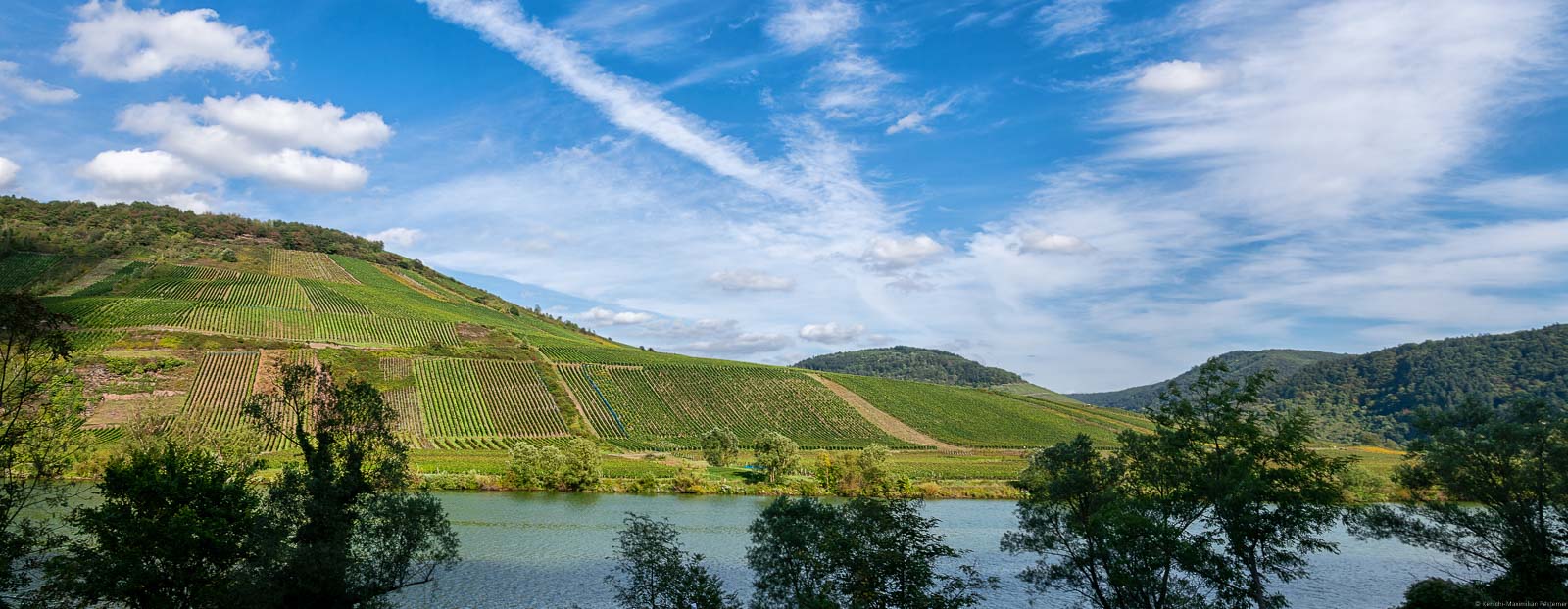 Vorne ist der Fluss Mosel. Dahinter liegt der Weinberg Briedeler Schäferlay. Oben ist Himmel.