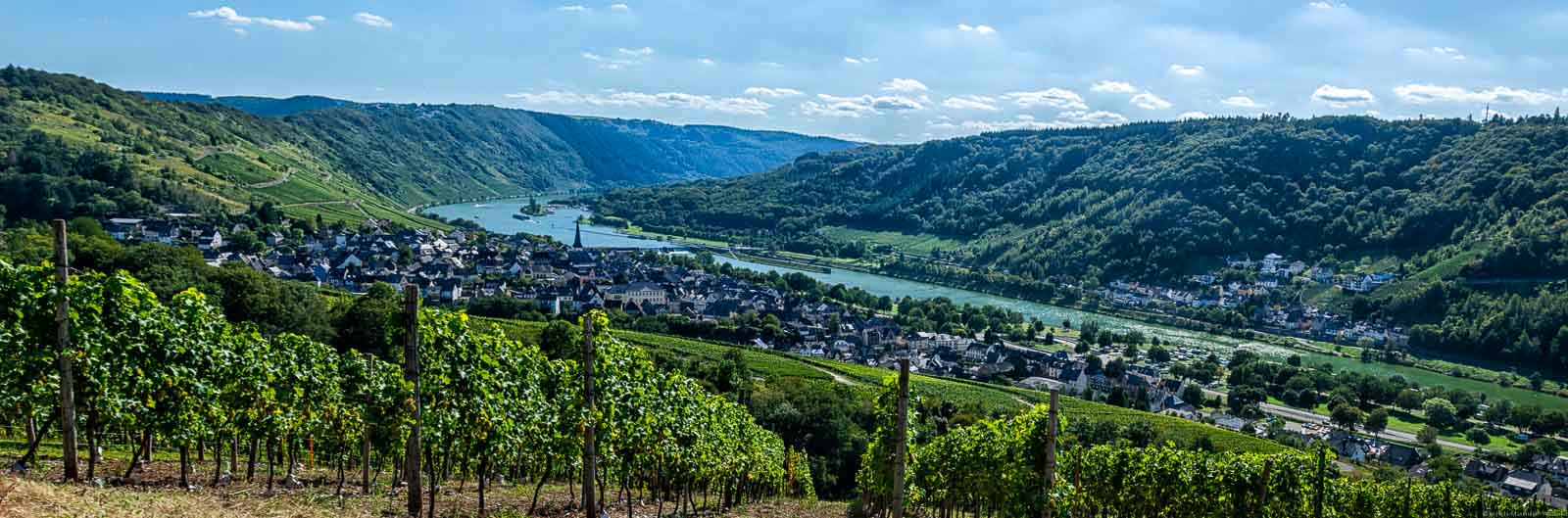 Bei den Orten Enkirch im Vordergrund und Kövenig im Hintergrund gibt es ein tolles Moselpanorama hinter dem Weinberg Enkircher Monteneubel. Zwischen den Orten fließt die Mosel. Im Hintergrund erkennt man weitere Weinberge und bewaldete Hänge. Der Himmel ist blau mit einigen Wolken.