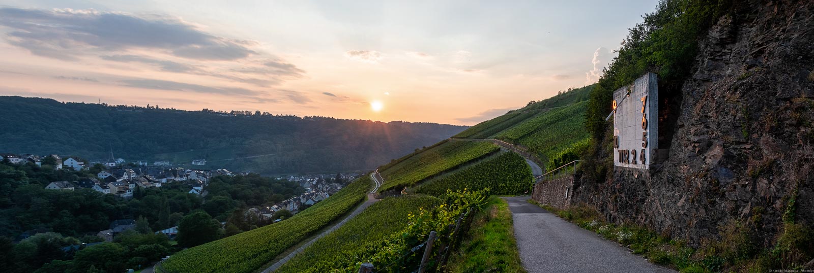 Vom Enkircher Steffensberg beim Ort Enkirch kann man tolle Sonnenuntergänge genießen. Vorne sind Weinberge. Rechts verläuft ein Weg unterhalb eines Felsens mit einer Sonnenuhr. Unten links ist ein Dorf. Im Hingergrund sind die Sonne, der Himmel und Wolken.