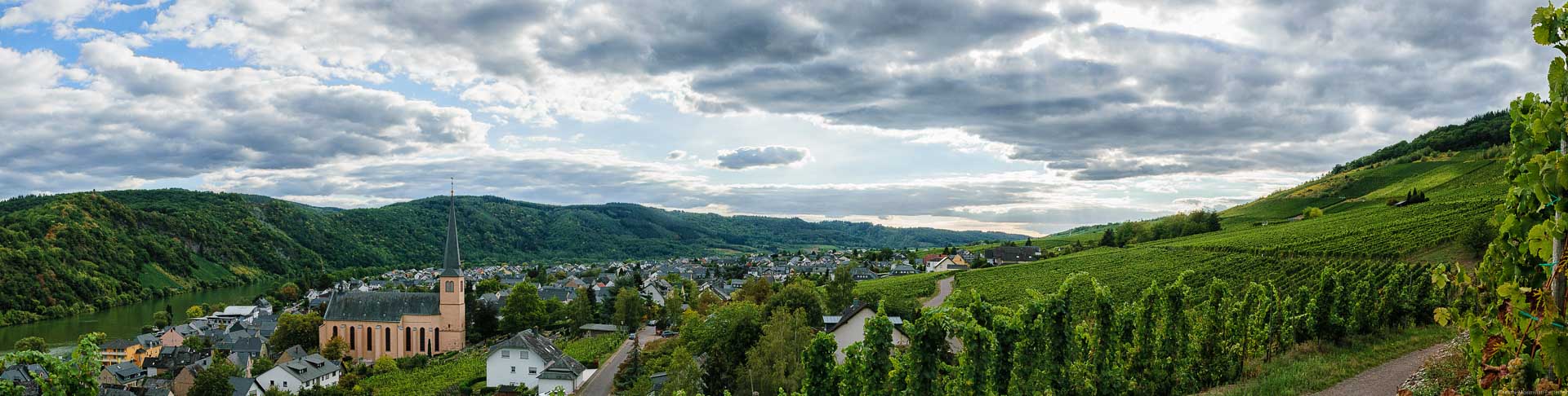 Die Kröver Kirchlay ist ein Weinberg nahe der Mosel an einem Hang. Vorne links fließt die Mosel. Am rechten Ufer liegt der Ort Kröv. Man sieht die Kirche des Ortes. Weiter rechts liegt der Weinberg. Am linken Bildrand und am linken Ufer der Mosel erkennt man den Weinberg Kröver Paradies. Am linken Ufer sind Wälder. Die blaue Himmel ist stark bewölkt.