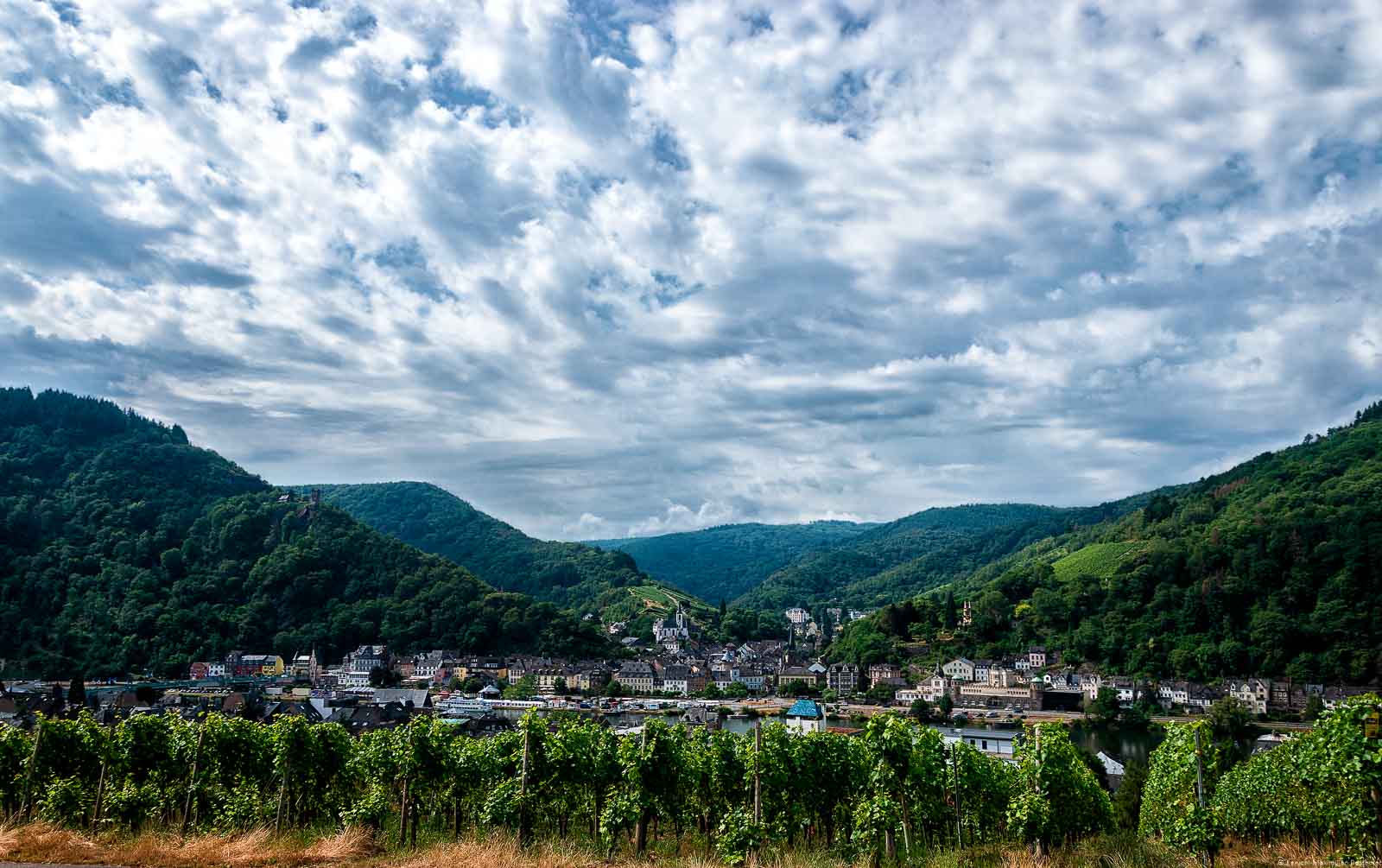 Die Altstadt von Traben-Trarbach ist geprägt vom Jugendstil. Im Hintergrund erkennt man den Trarbacher Burgberg und den Trarbacher Schlossberg​.Die Mosel fließt durch den Ort. Der Himmel ist stark bewölkt. Die Doppelstadt liegt im Bereich der Mittelmosel.