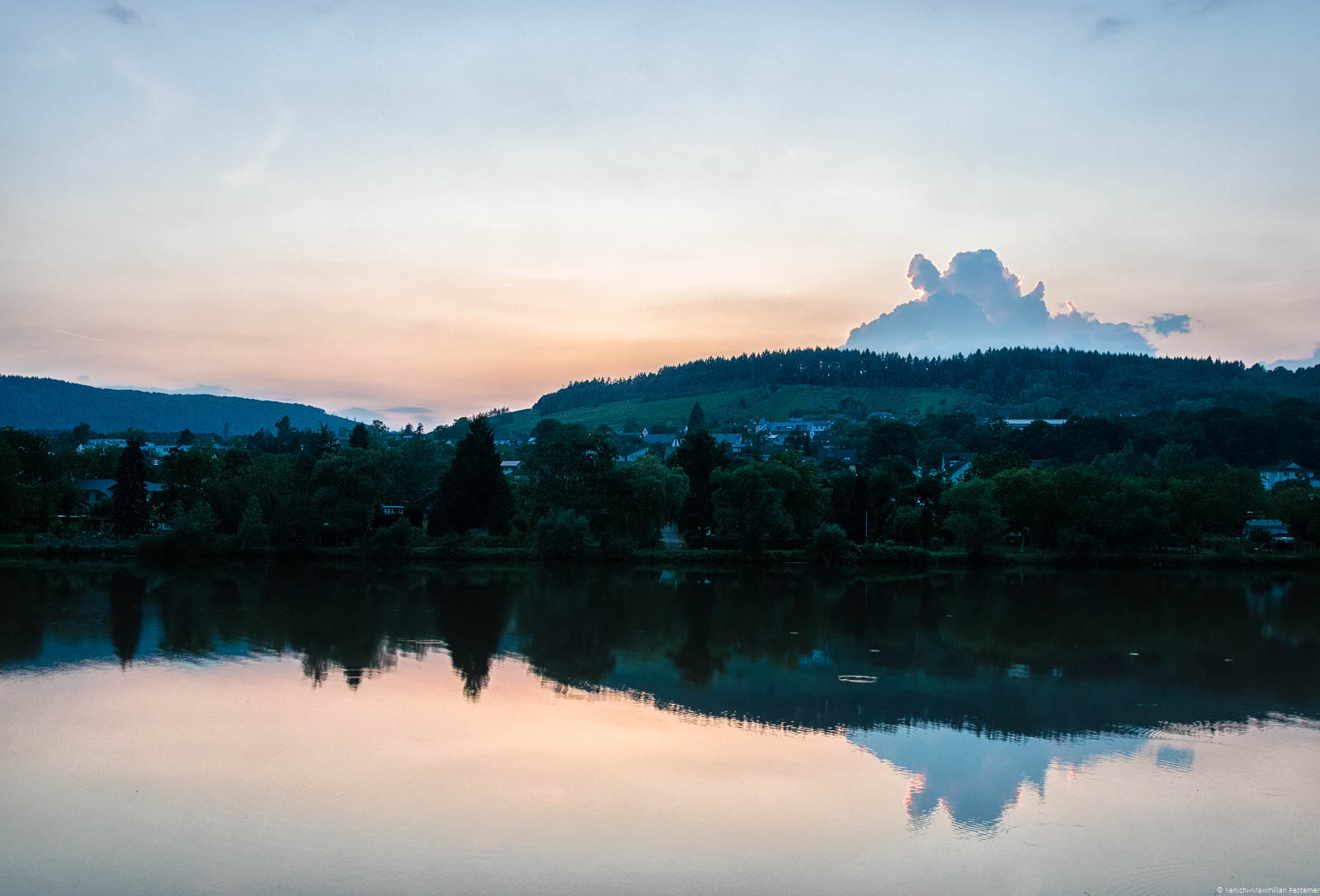 Vorne fließt die Mosel. In dem Fluss Mosel spiegeln sich das Ufer, die Bäume und der Weinberg im Hintergrund. Die Sonne geht am Himmel unter, sodass der Himmel in orange und blau gefärbt ist. Man erkennt außerdem einige Häuser.