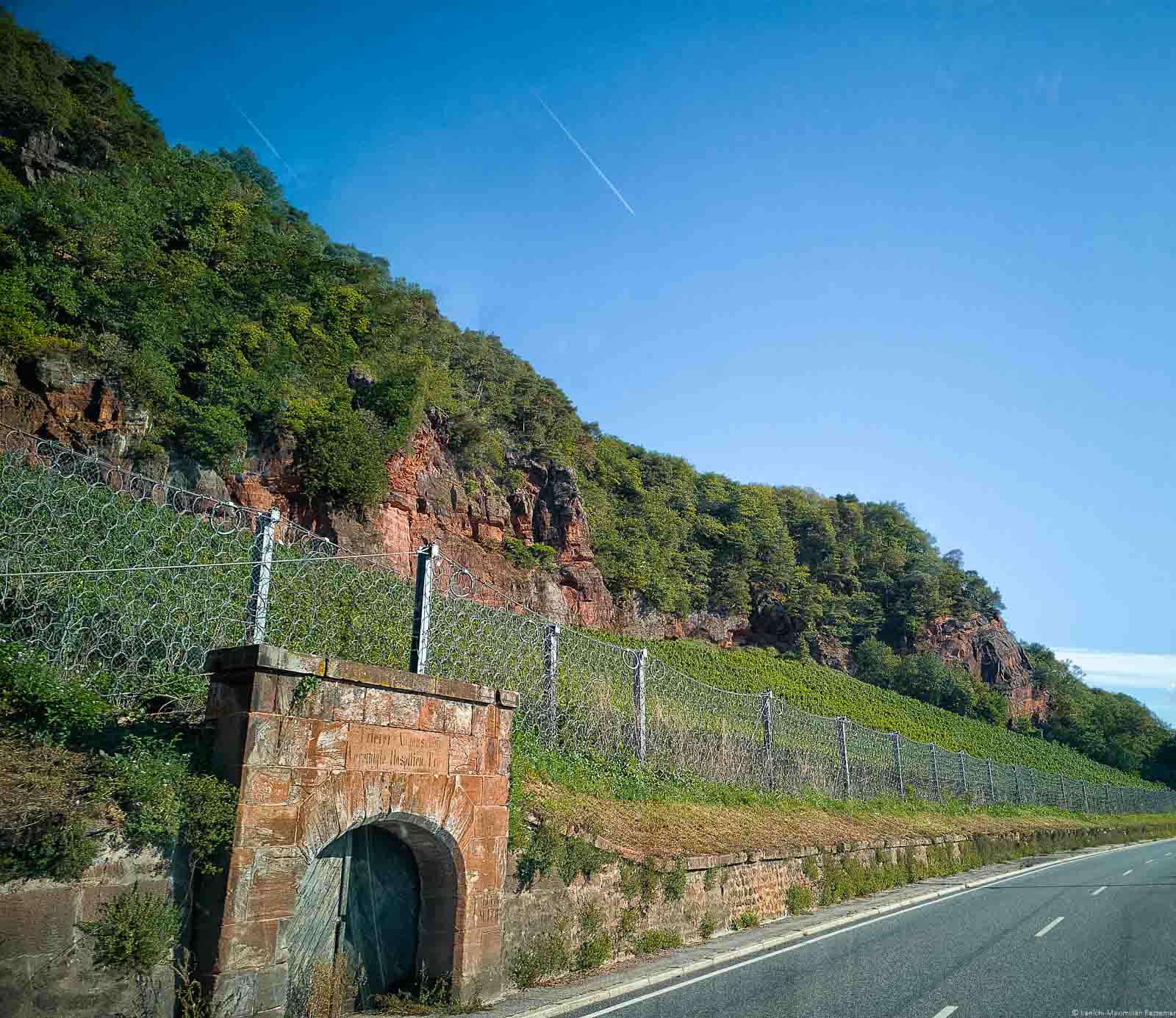 Vorne rechts befindet sich eine Straße. Auf der linken Seite ist der Weinberg Trierer Augenscheiner. Weiter hinten links sind bewaldete rote Felsen aus Buntsandstein. Oben ist der Himmel blau ohne Wolken.