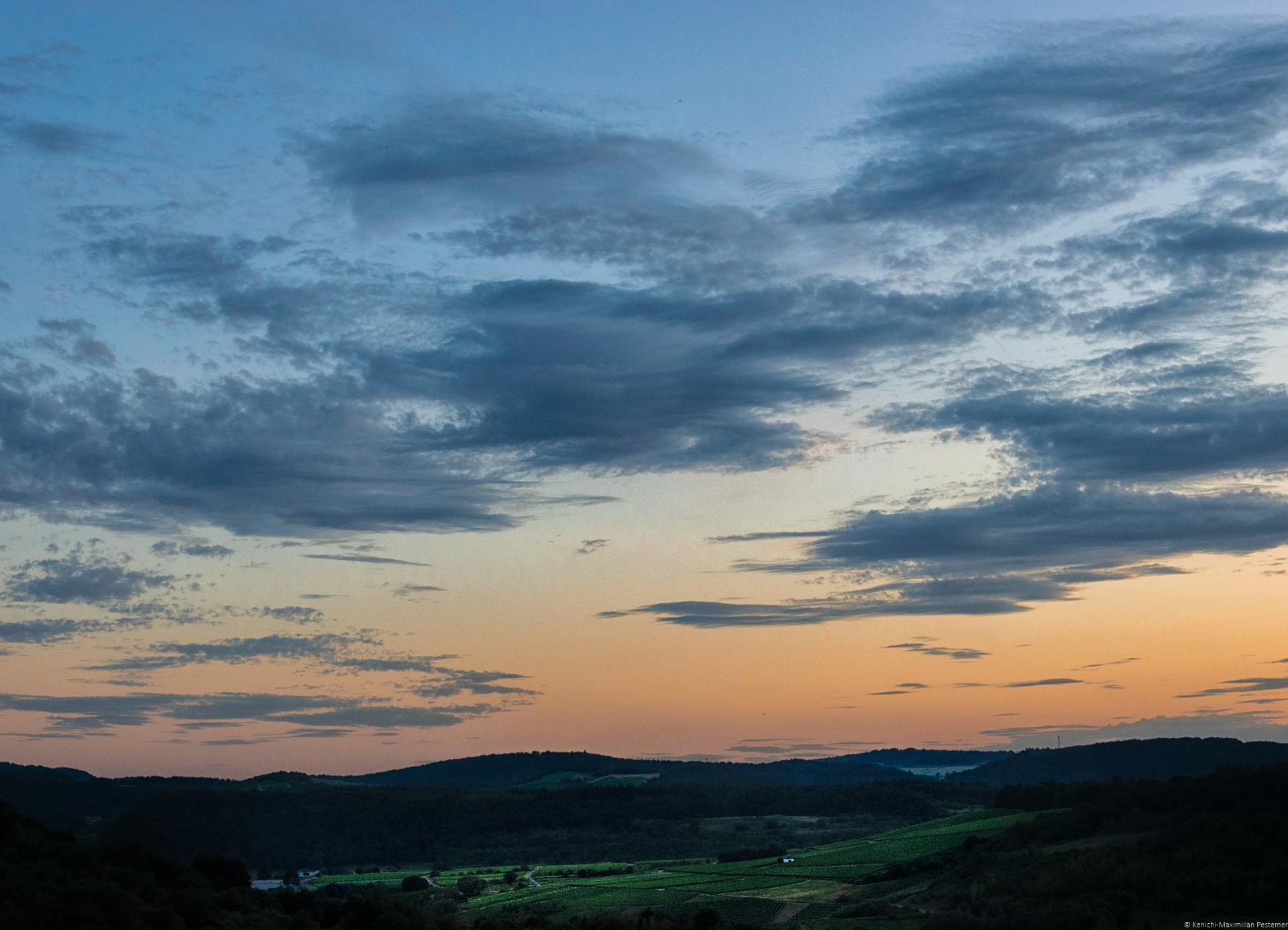 Man blickt aus der Ferne auf den Weinberg Wiltinger Schlangengraben. Rund um den Weinberg erkennt man bewaldete Hügel. Im Weinberg sind einige kleine Häuser. Links im Bild befindet sich der Fluss Saar. Am Himmel geht die Sonne unter. Der Himmel ist orange-rot und blau. Es sind Wolken am Himmel.