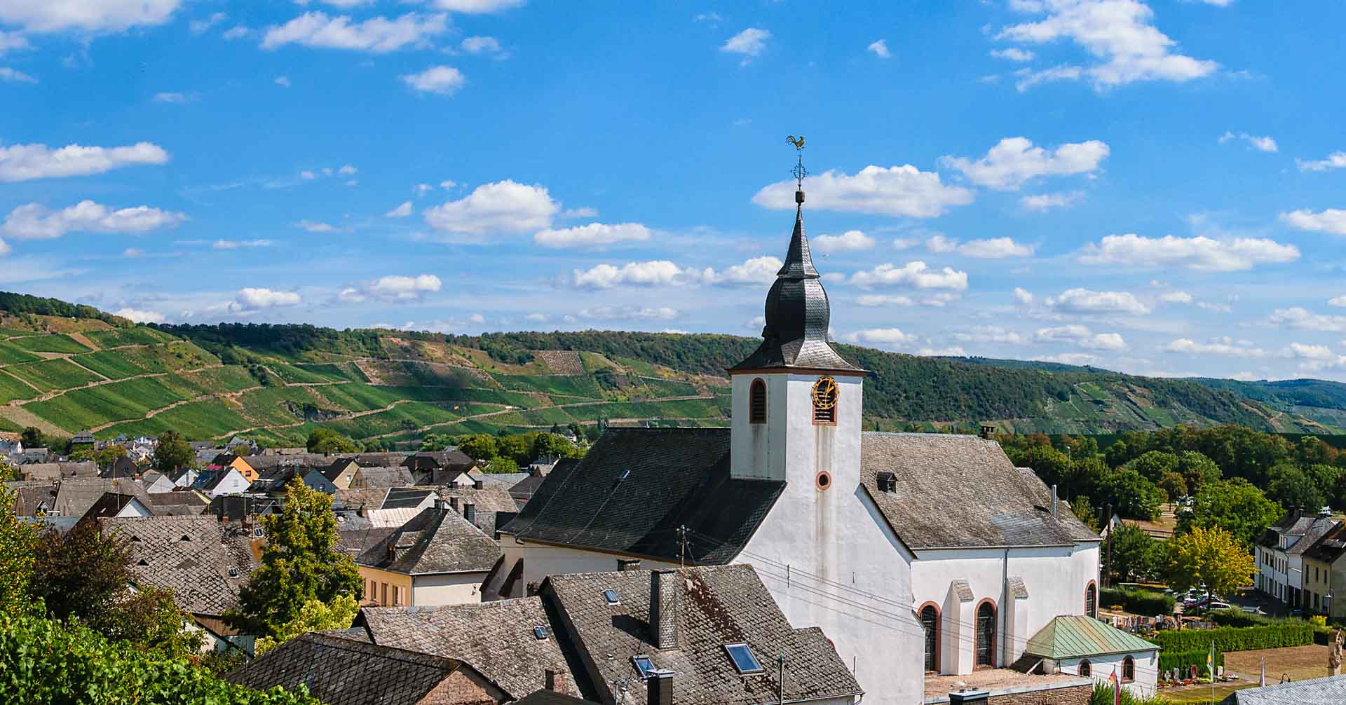 Man erkennt die Kirche des Ortes Klüsserath an der Mosel und den Weinberge Klüsserather Bruderschaft im Hintergrund. Es sind weitere Häuser erkennbar. Der blaue Himmel ist ganz leicht bewölkt.