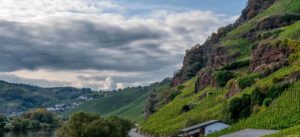 Der steile Weinberg Erdener Prälat befindet sich auf der rechten Seite des Bildes. Vor dem Weinberg verläuft eine Straße. Man erkennt auch das Gebäude der alten römischen Kelteranlage. Links im Bild fließt die Mosel mit Bäumen an der Uferrändern. Im Hintergrund links liegt der Ort Ürzig am Himmel sind große Wolken.