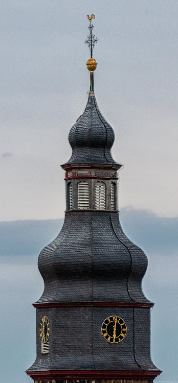 Man erkennt den Kirchturm des Ort Kallstadt in der Weinregion Pfalz. Am Kirchturm befindet sich eine Uhr. Im Hintergrund ist eine dunkle Wolke am Himmel.