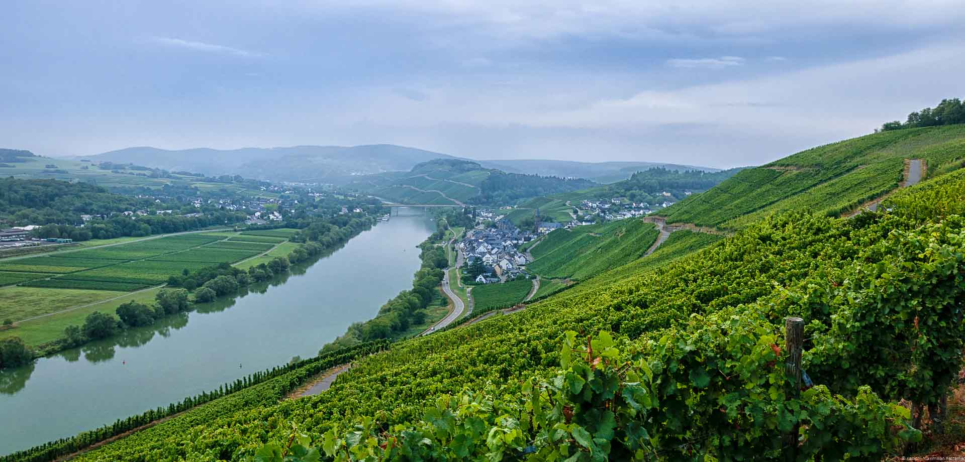 Man blickt auf das Moseltal mit Nebel im Hintergrund. Am rechten Ufer der Mosel liegt vorne der steile Hang des Weinbergs Lieserer Niederberg-Helden. Dahinter liegt das Dorf Lieser. Im Hintergrund sind Weinberge und bewaldete Hügel. Der Himmel ist stark bewölkt.