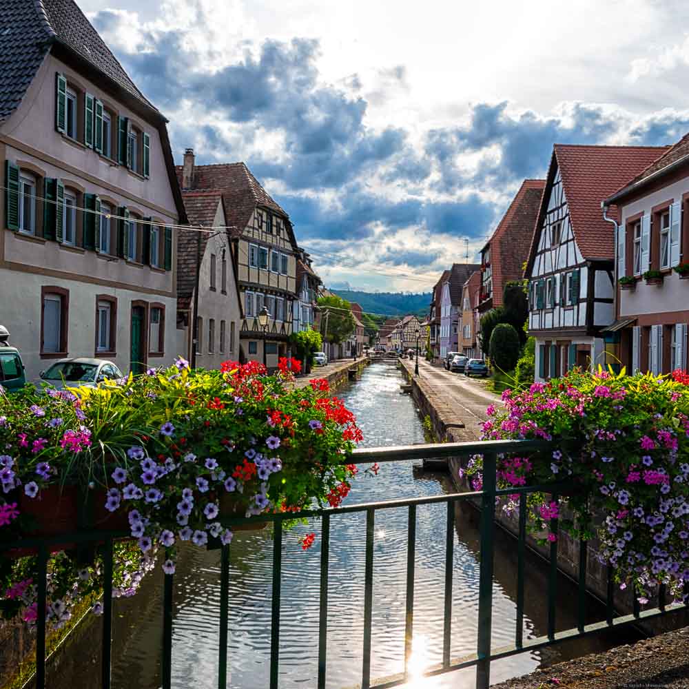 Brücke mit Blumenkästen über den Lauter in Weißenburg Elsass. An den Ufern des Flusses Lauter befinden sich Häuser. Der Himmel ist bewölkt.