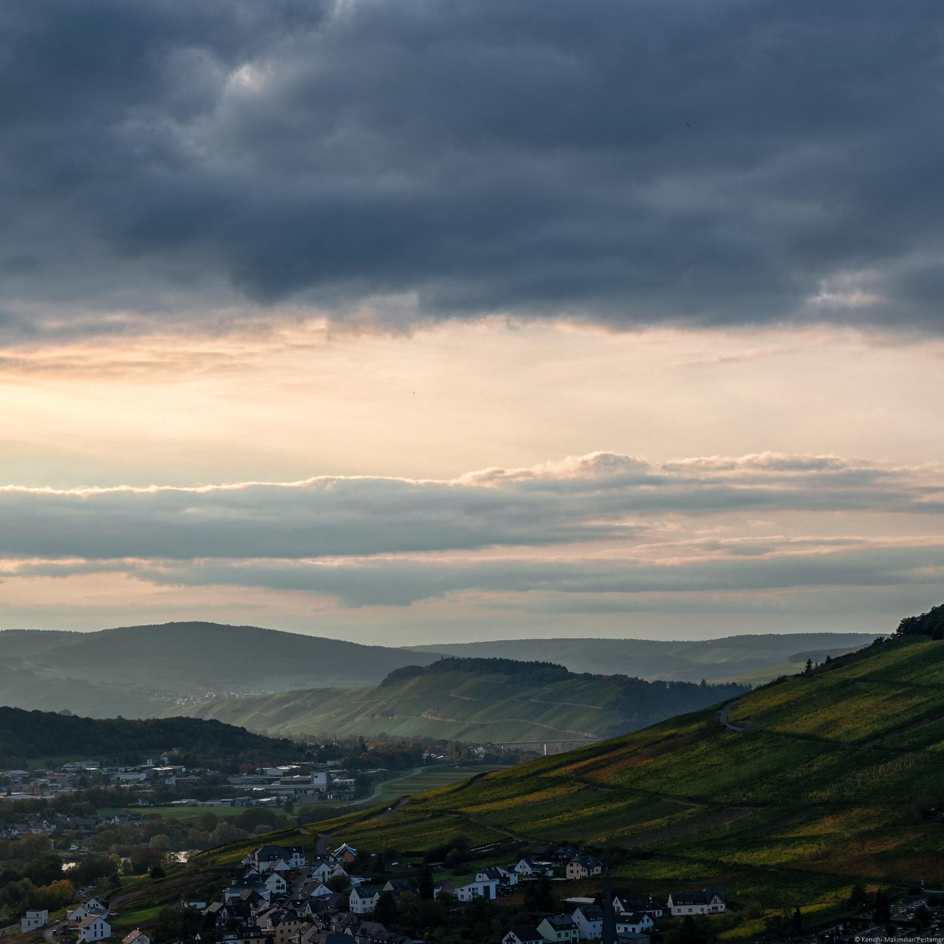 Im Vordergrund links befindet sich Bernkastel-Kues. Dahinter rechts liegt der Weinberg Kueser Weisenstein. Etwas weiter links liegt der Ortsteil Andel. Dahinter liegt der Weinberg Brauneberger Juffer Sonnenuhr. Es folgen weitere Weinberge und bewaldete Hügel. Der Himmel ist gelb-orange gefärbt. Oben erkennt man eine dunkel-blaue Wolke.