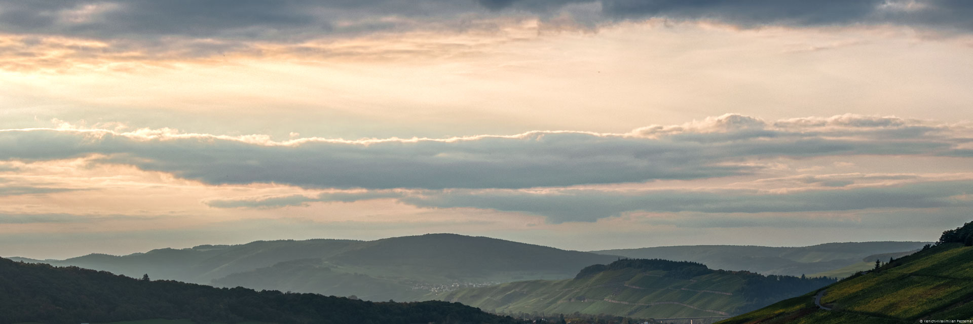 In der Mitte erkennt man den Weinberg Brauneberger Juffer Sonnenuhr am späten Abend. Links und rechts sind weitere Weinberge und bewaldete Hügel. Am Himmel sind blau-graue Wolken und eine orangene Färbung erkennbar.
