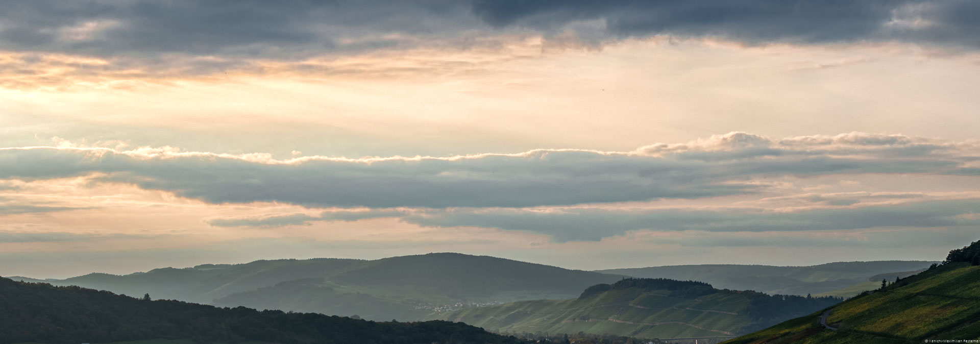In der Mitte erkennt man den Weinberg Brauneberger Juffer Sonnenuhr am späten Abend. Links und rechts sind weitere Weinberge und bewaldete Hügel. Am Himmel sind blau-graue Wolken und eine orangene Färbung erkennbar.