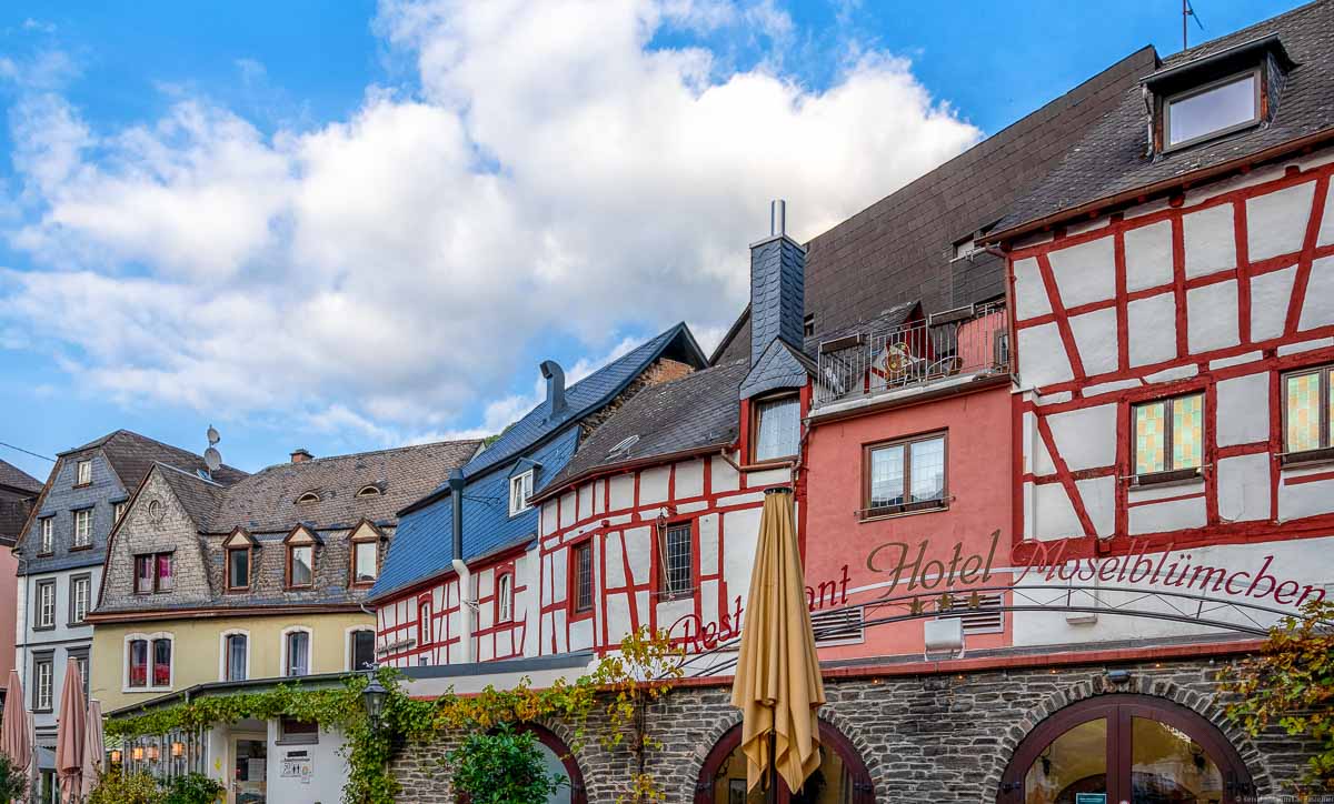 Auf dem Foto sind Häuser mit Fachwerk-Fassaden am Karlsbader Platz in Bernkastel-Kues. Am blauen Himmel sind in der Mitte oben und links oben einige Wolken.