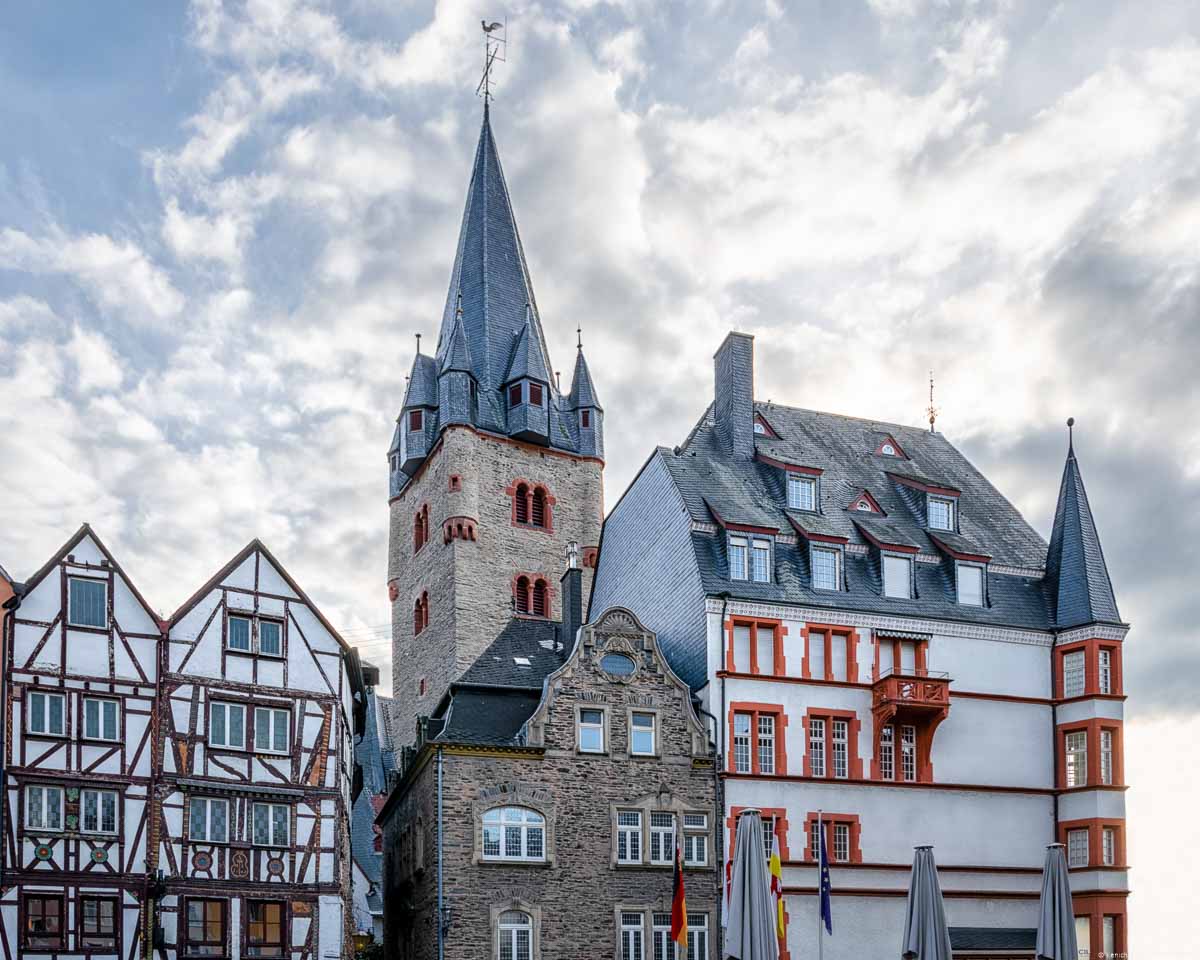 Vorne links ist ein Haus mit Fachwerk-Fassade. In der Mitte ist ein Haus mit Schiefer-Mauern. Rechts ist ein größeres weißes Gebäude mit roten Fensterrahmen. Das Dach ist aus blauem Schiefer. Dahinter ragt der Turm der Pfarrkirche St. Michael in den blauen und bewölkten Himmel von Bernkastel-Kues empor.