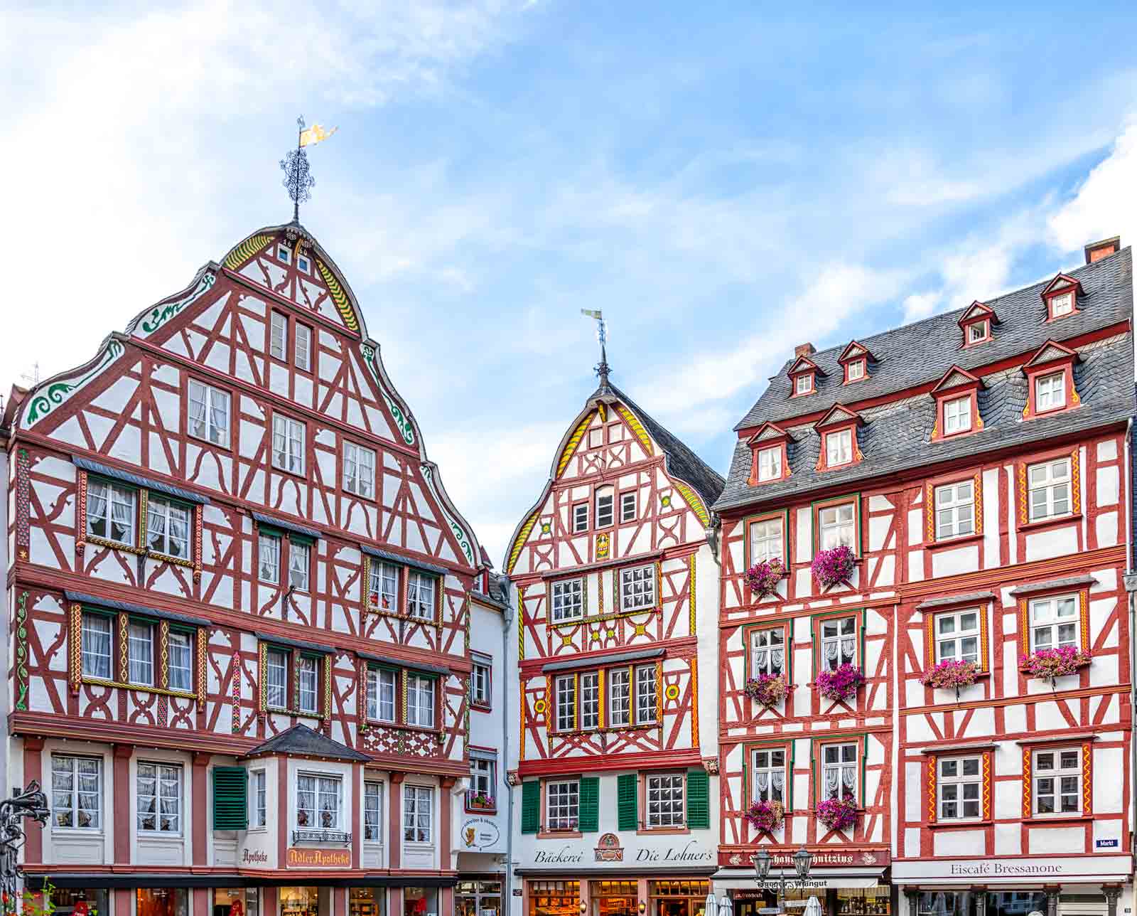 Man erblickt drei Häuser mit Fachwerk-Fassaden am Marktplatz von Bernkastel-Kues. Unten sind Läden. Am blauen Himmel sind Wolkenschlieren.