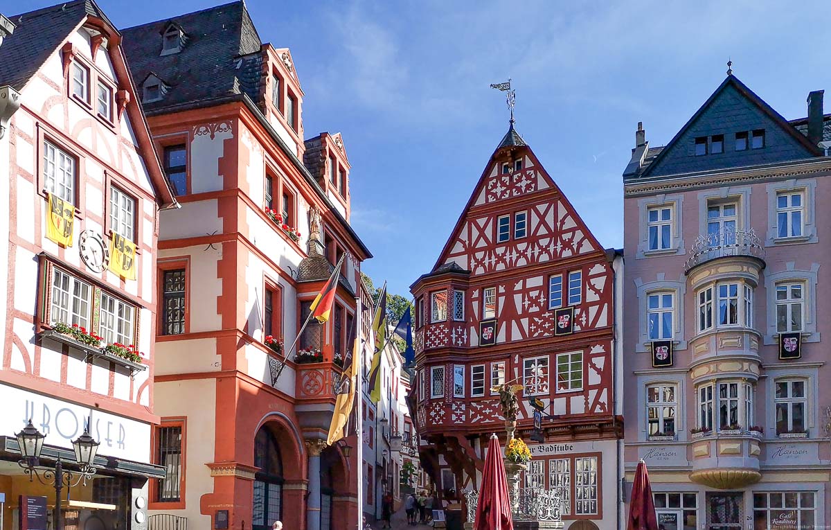 Man sieht Häuser am Marktplatz von Bernkastel-Kues rund um den Michaelsbrunnen. Die Häuser haben Fachwerk-Fassaden. Oben ist noch etwas blauer Himmel.
