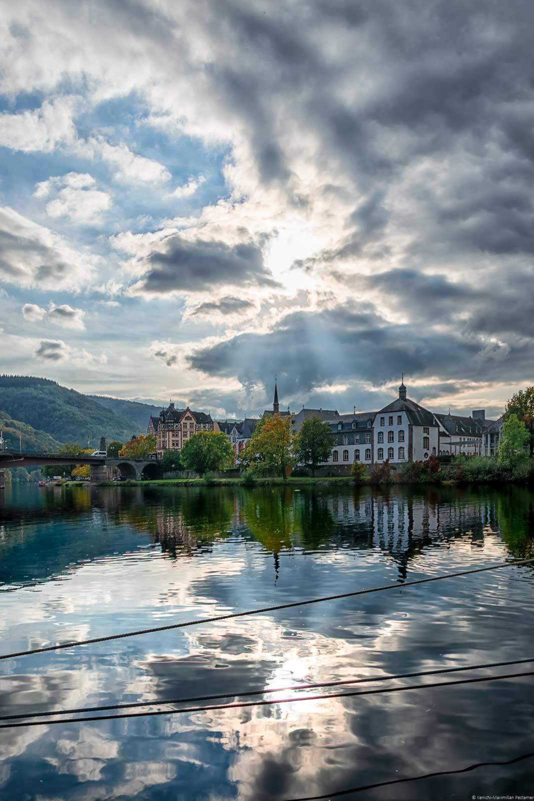 Im Vordergrund spiegelt sich das St. Nikolaus Hospital. Dieses ist auch als Cusanusstift bekannt und befindet sich am Ufer der Mosel in Bernkastel-Kues. Daneben erkennt man links in der Mitte eine Brücke und weitere Gebäude. Am hellen Himmel strahlt die Sonne durch die großen Wolken hindurch.