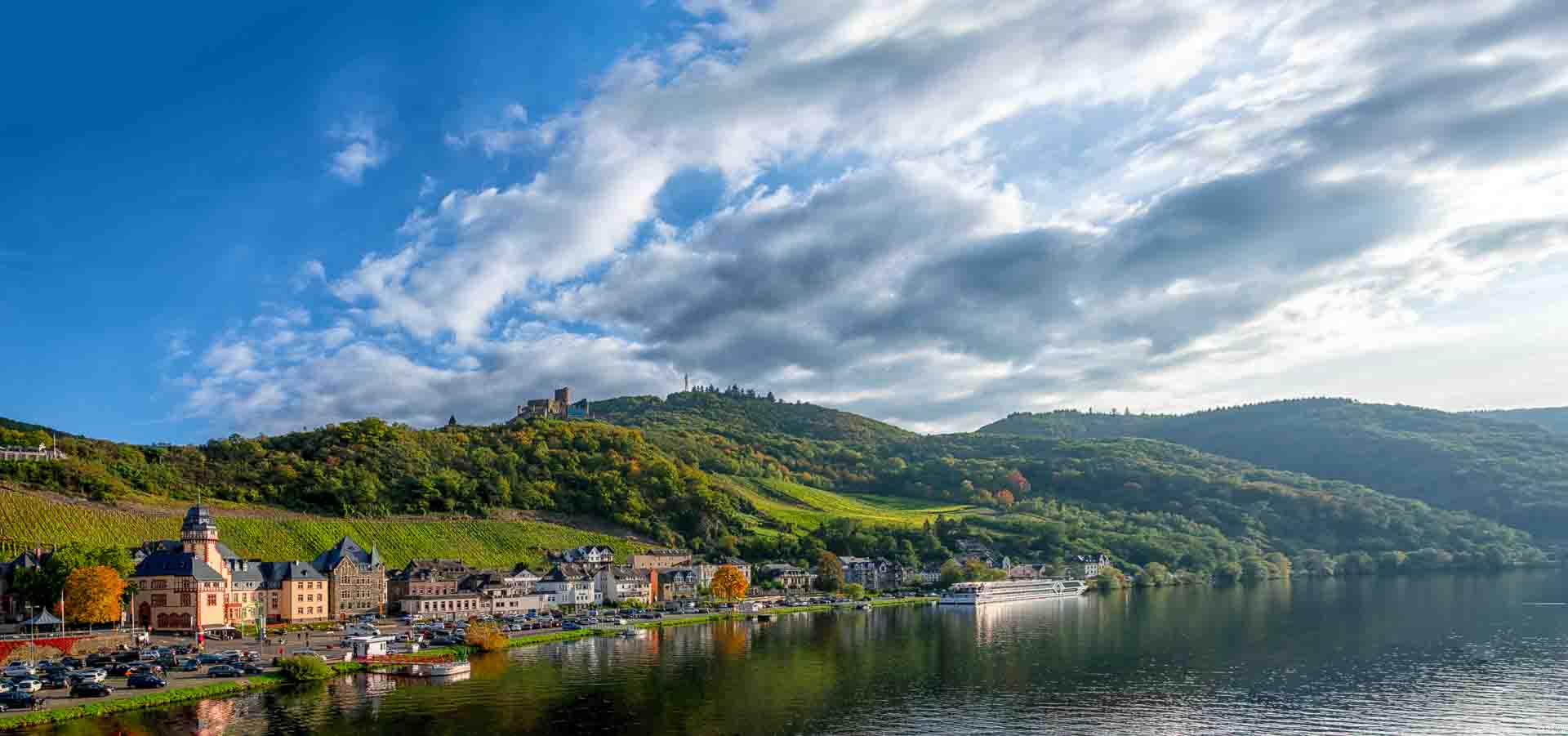 Im Vordergrund rechts fließt die Mosel. In der Mosel spiegelt sich die Uferpromenade. Am linken Ufer erkennt man Häuser des Ortes Bernkastel-Kues. Aa Hang hinter der Uferpromenade links befindet sich der Weinberg Bernkasteler Schlossberg zusammen mit Wälder. Oben auf dem Hang befindet sich die Burgruine Grevenburg. Am blauen Himmel sind viele Wolken.
