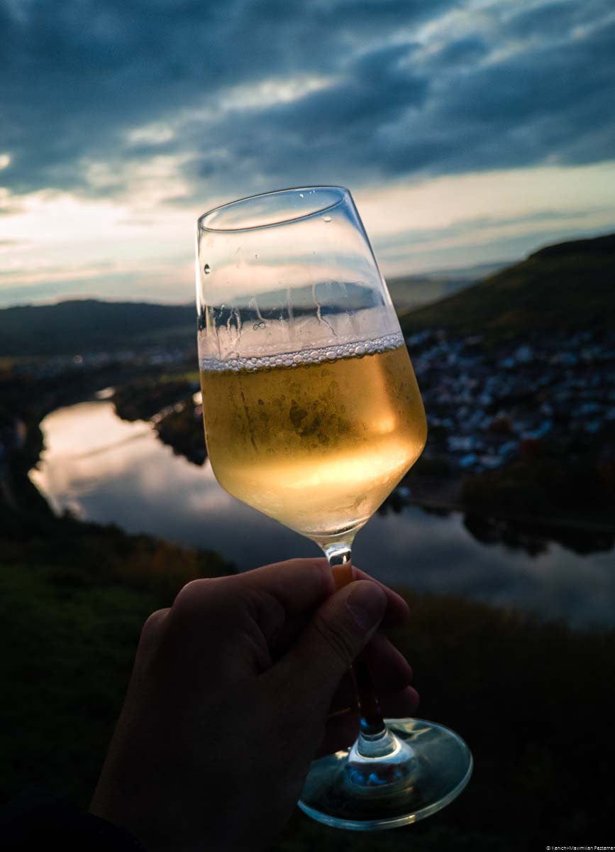 Am Abend wird ein Glas Riesling im Vordergrund genossen. Im Hintergrund befinden sich die Mosel, der Weinberg Kueser Weisenstein, weitere Weinberg und ein atemberaubender Sonnenuntergang.