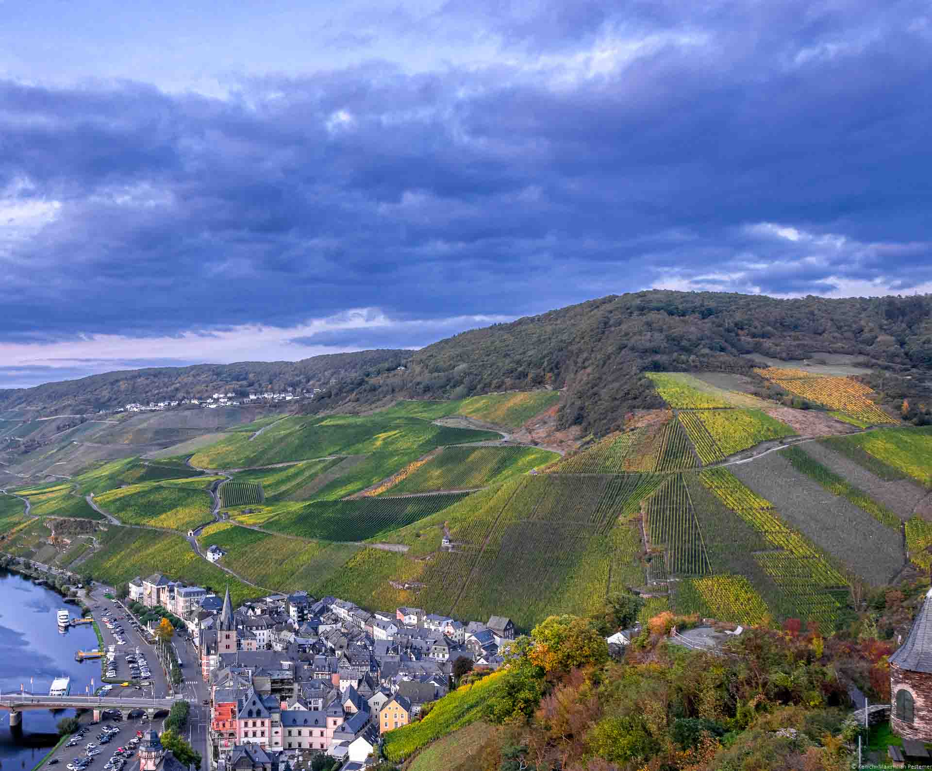 Man sieht den Ort Bernkastel-Kues am Ufer der Mosel unten links im Bild. An der rechten Seite liegt der Weinberg Alte Badstube am Doctorberg. Auf dem Bild ist ein umfassende Weinbergslandschaft am späten Abend erkennbar. Am Himmel sind lila Wolken.