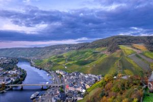 Am Abend blickt man auf den Ort Bernkastel-Kues, die Mosel und die dahinter liegenden Weinberge. Am Himmel sind lila Wolken.
