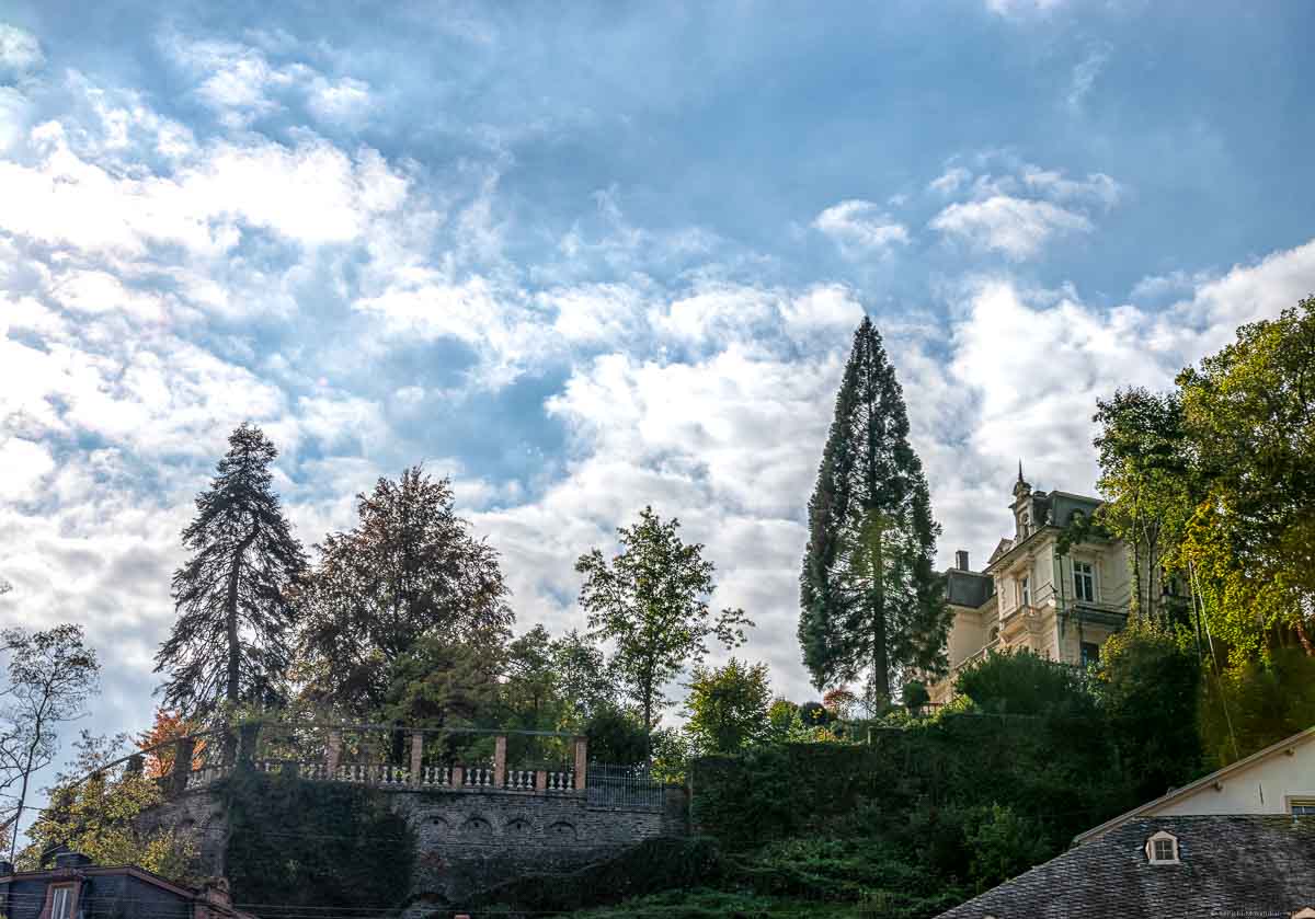 Auf einem bewaldeten Berg ist eine Villa in gelber Farbe im Ort Traben-Trarbach. Am blauen Himmel strahlt die Sonne durch Wolken.