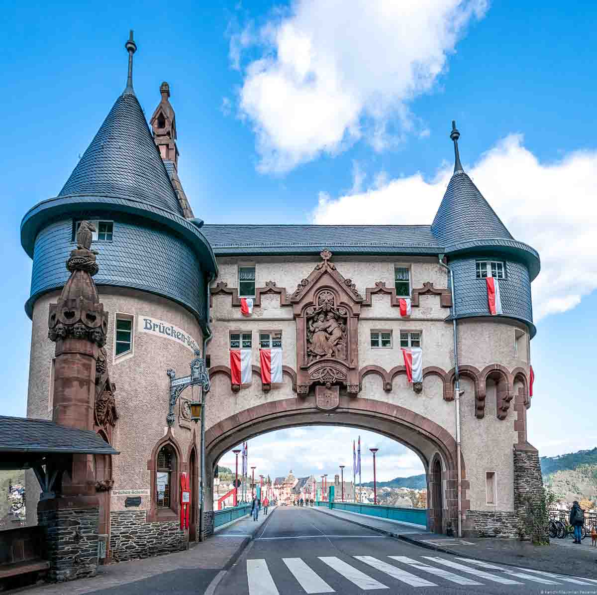 Im Vordergrund führt eine Straße mit Zebrastreifen unter dem Trarbacher Brückentor hindurch. Am blauen Himmel erkennt man mindestens zwei größere Wolken.
