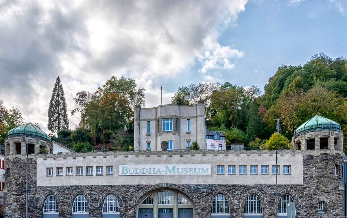 Man erkennt ein Gebäude aus Schiefersteinen, welches drei Türme hat. Es ist das Buddha-Museum in Traben-Trarbach. Hinter dem Gebäude ist ein bewaldeter Hügel. Am blauen Himmel ist eine große Wolke links.
