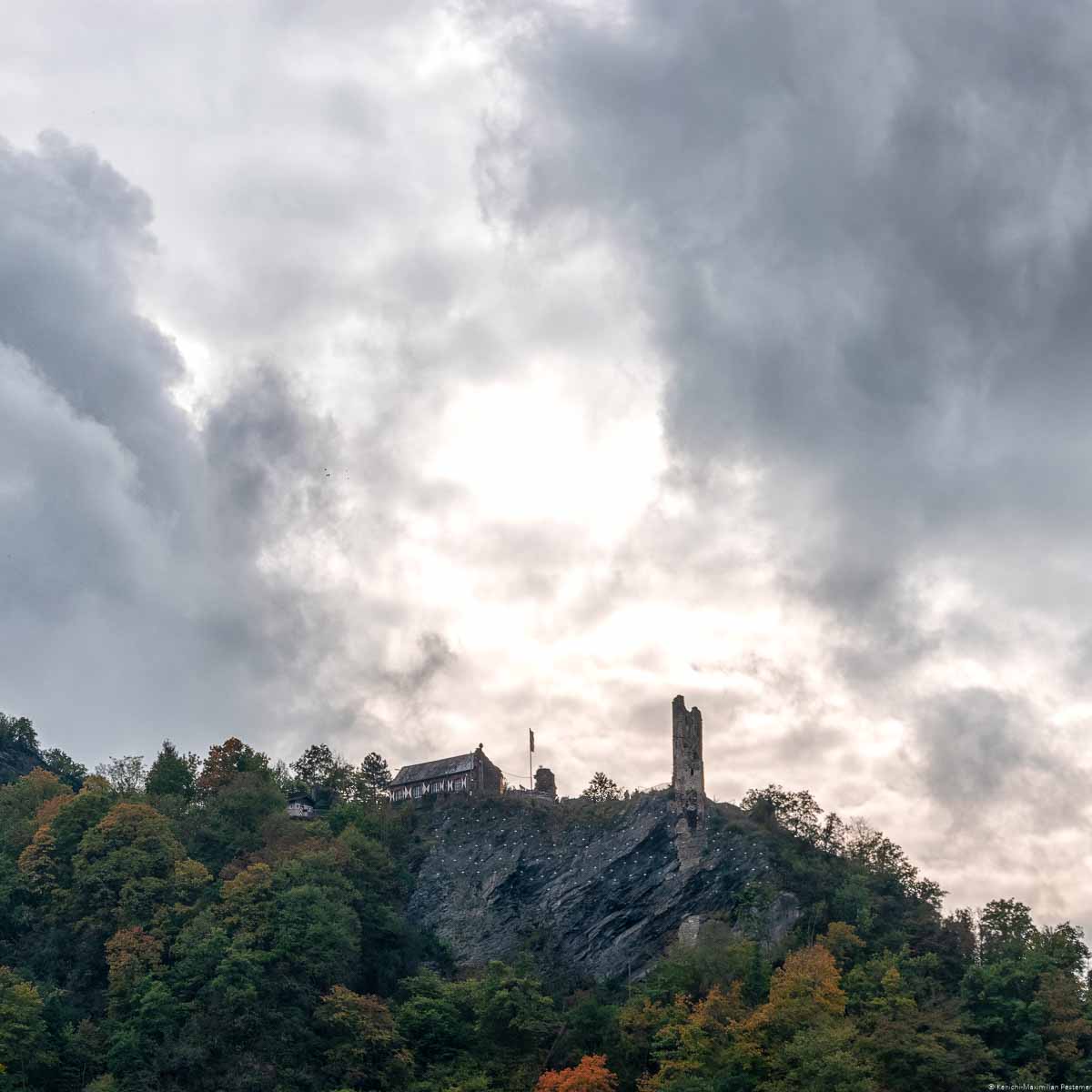 Auf einem steilen und felsigen Berg liegt die Grevenburg oberhalb von Traben-Trarbach. Der steile Berg ist mit Bäumen in herbstlicher Färbung bewachsen. Der Himmel oberhalb der Burg ist stark bewölkt.