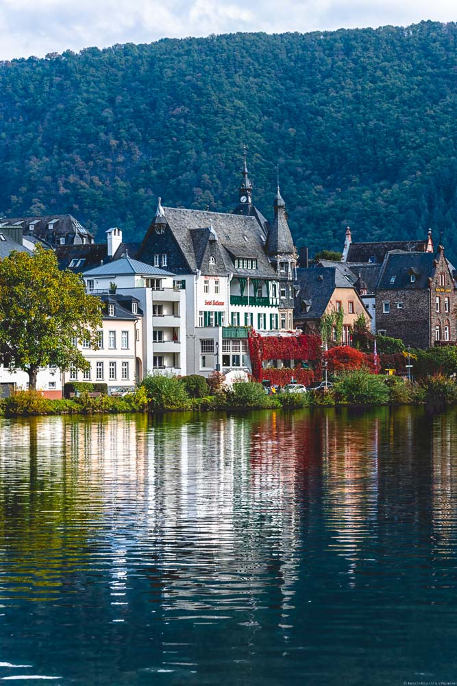 Man blickt auf die Mosel. In der Mosel spiegelt sich das Hotel Bellevue, welches im Jugendstil erbaut wurde. Daneben erkennt man weitere Gebäude am Ufer des Flusses. Im Hintergrund ist ein bewaldeter Hügel. Oben erkennt man einige Wolken.
